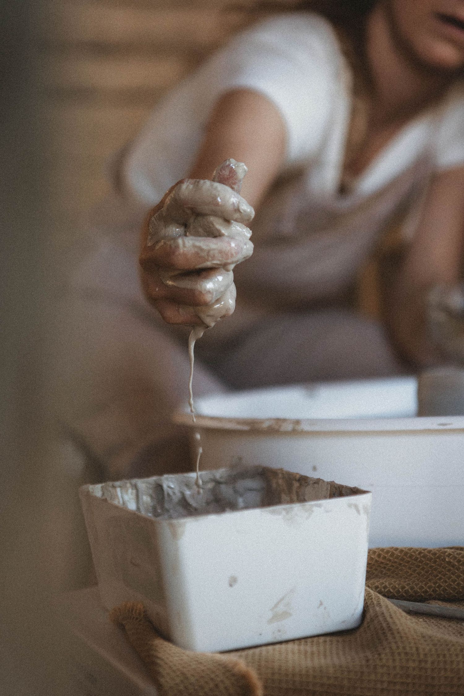 Clay-covered hands working with pottery materials over a white container.