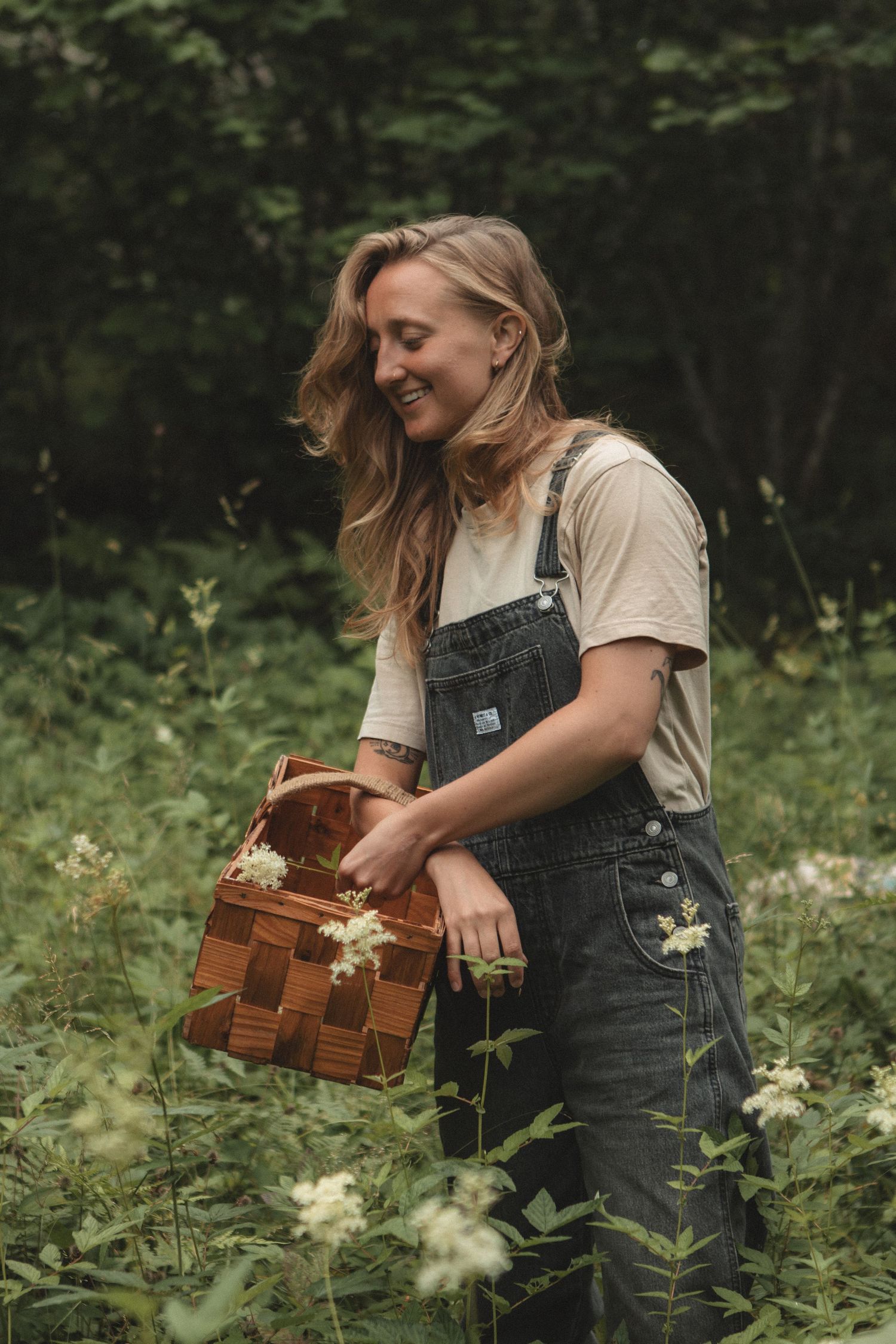 Person in overalls gathering wildflowers in forest with woven basket.