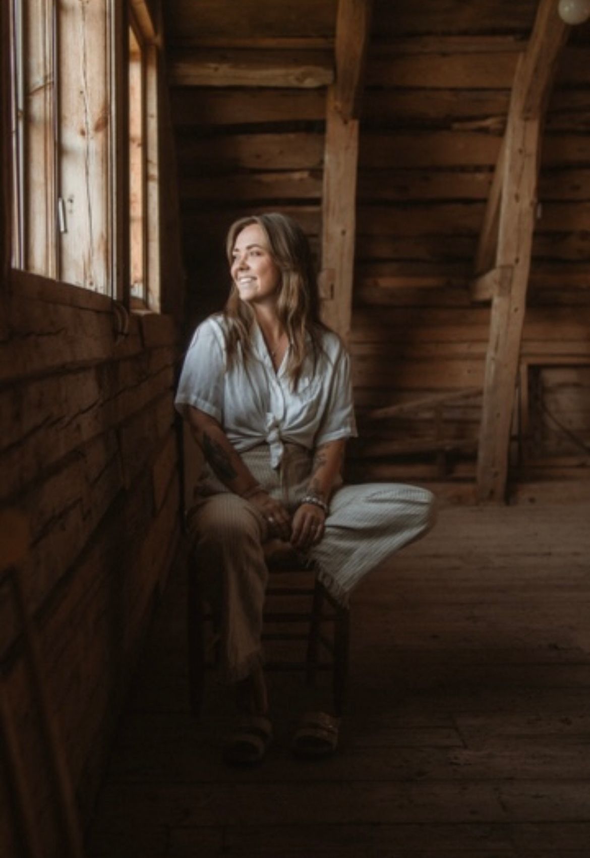 A person in casual attire sits by a rustic wooden window in a barn loft, gazing at natural light streaming in.