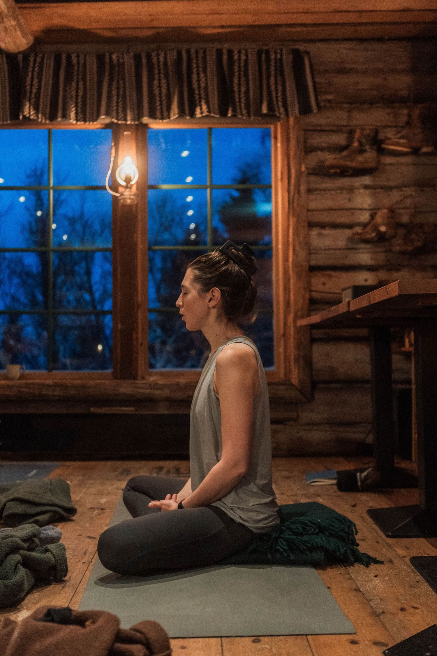 A person meditating on a yoga mat inside a cozy log cabin at dusk with a warm lantern glowing in the window.