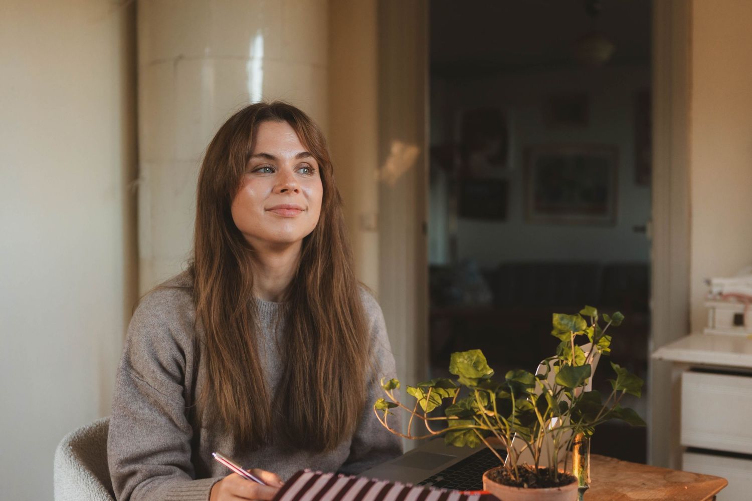 A woman in a cozy sweater sits at a table with a notebook and potted plant in warm indoor lighting.