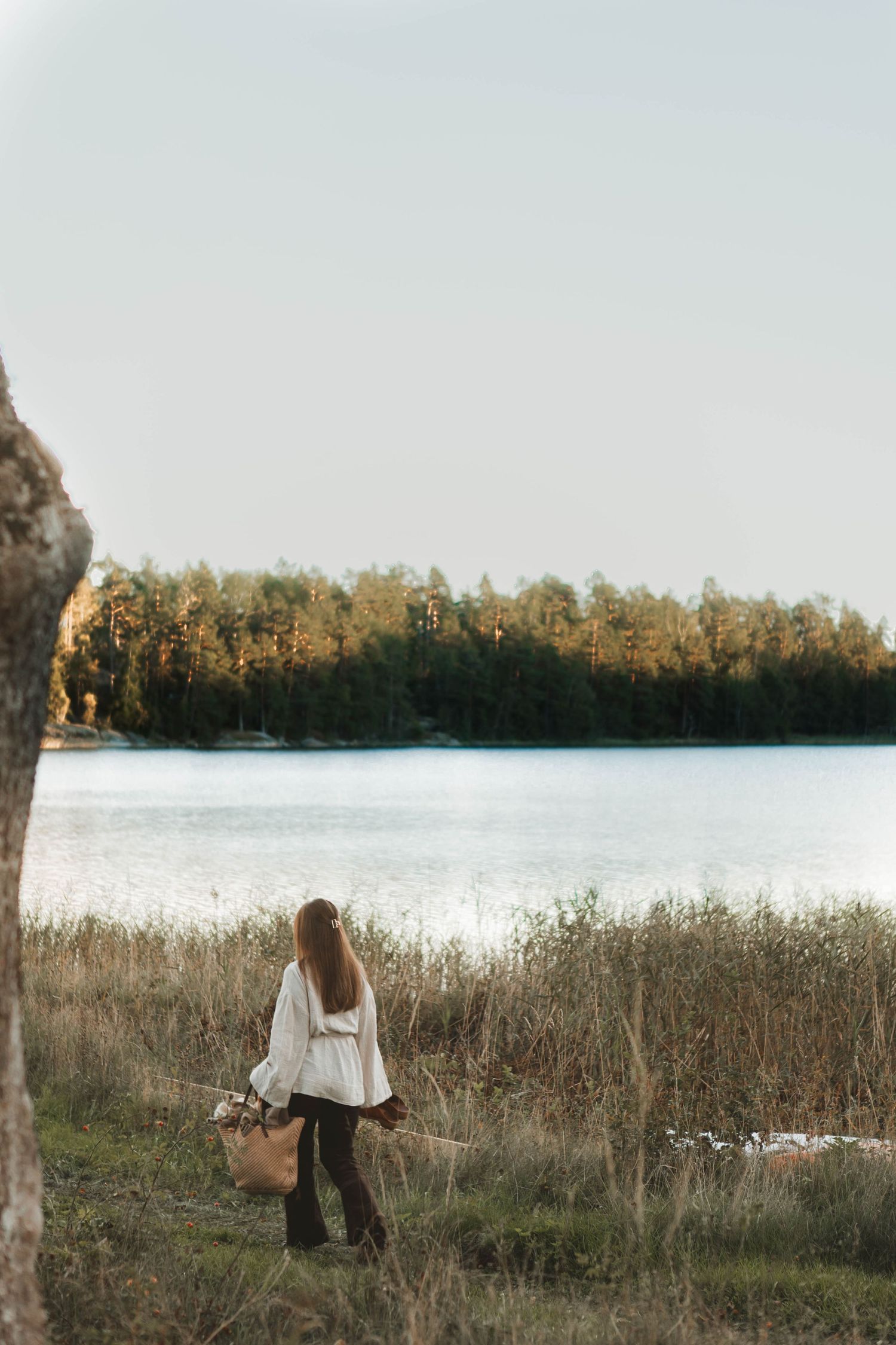Person in white sweater stands near a serene lake at sunset with tall trees in the background.