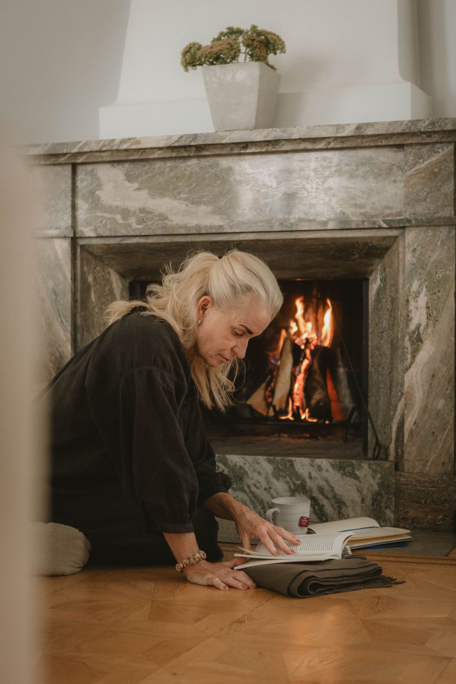 Person reading book by warm fireplace in relaxing home environment.