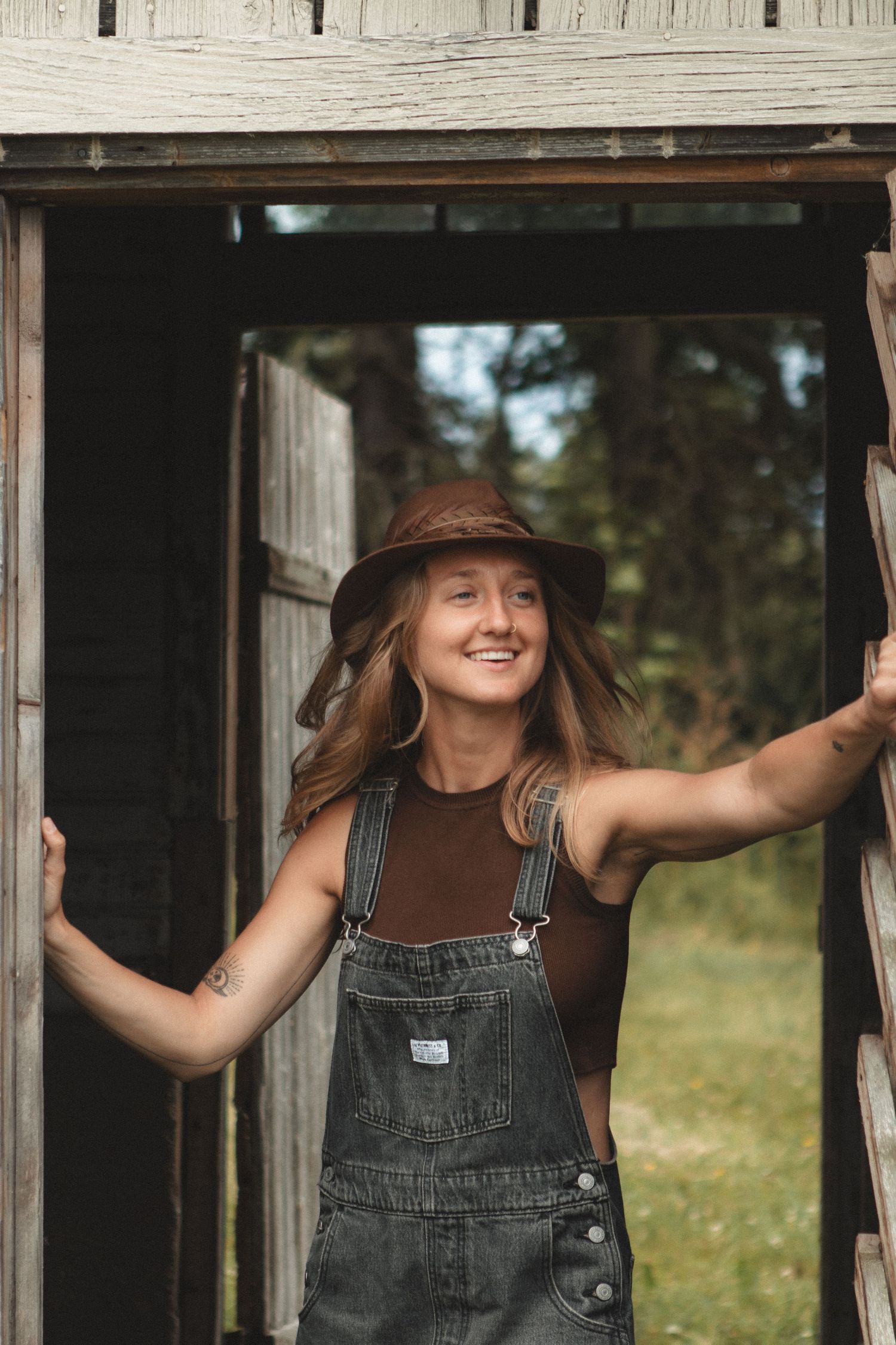 Person in overalls and brown hat standing in rustic doorway of wooden building.