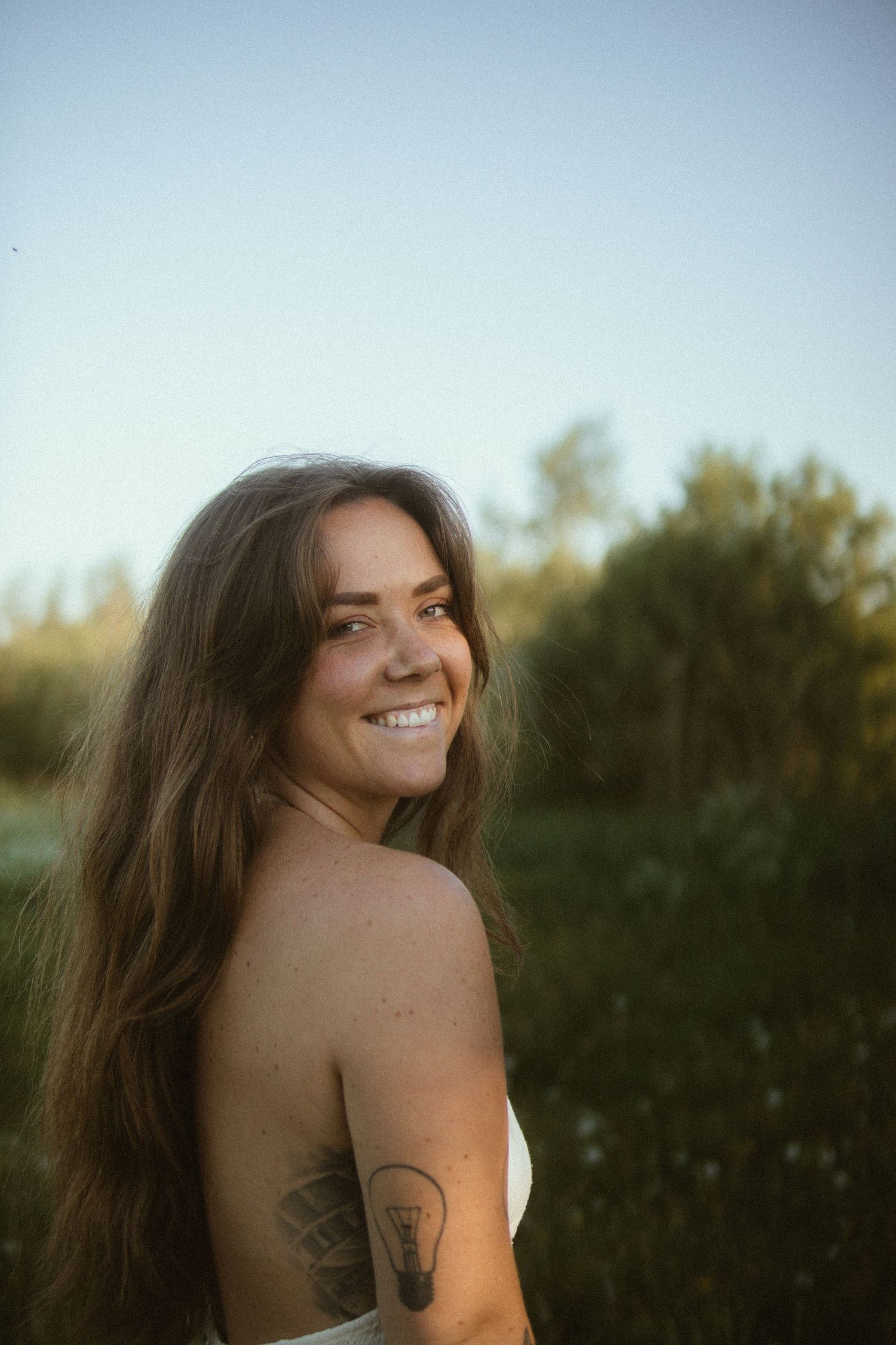 A person with long dark hair and a tattoo smiles while posing in nature during sunset.
