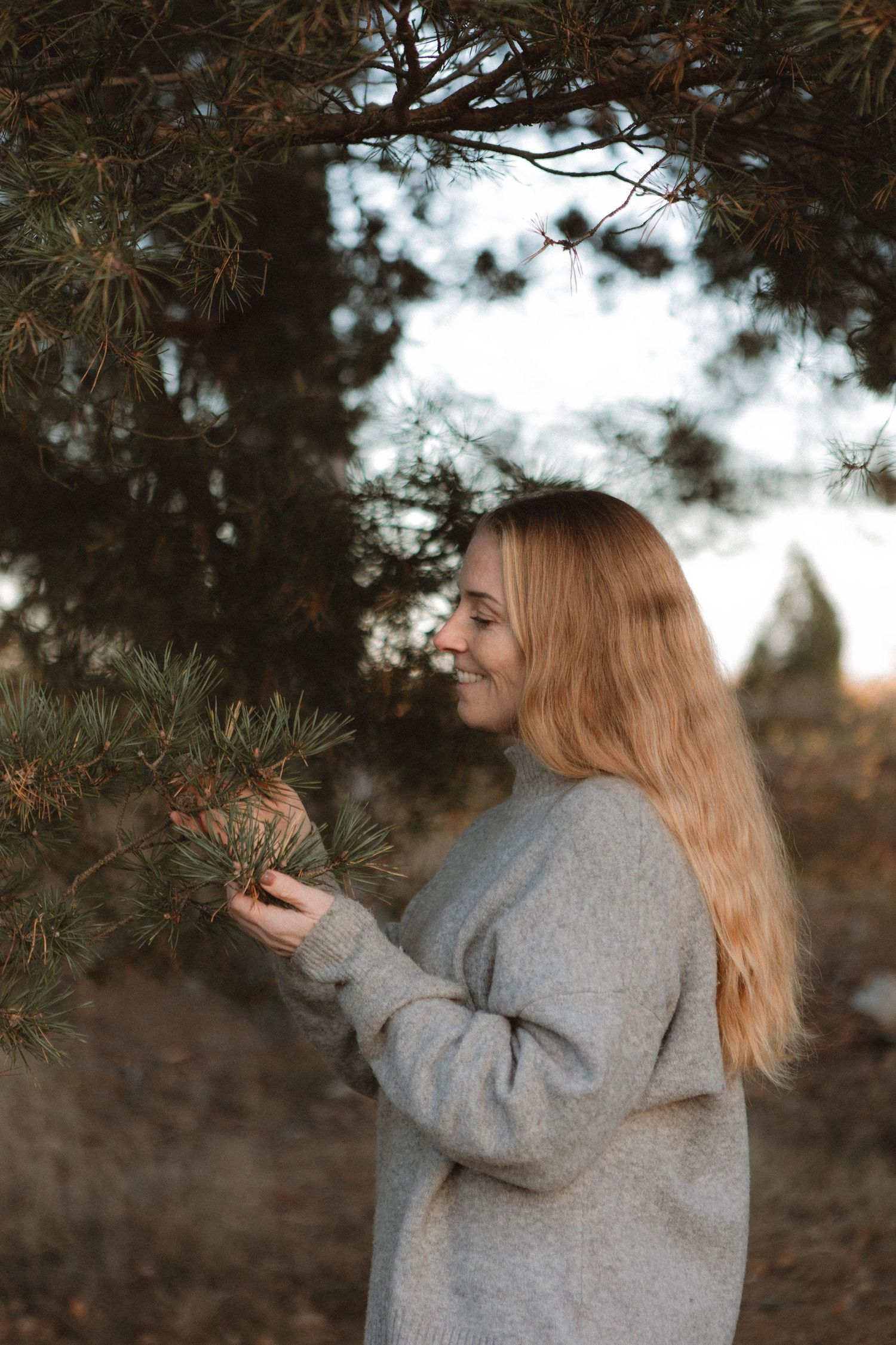 Woman in a cozy gray sweater smiles while touching pine branches on an autumn day in nature.