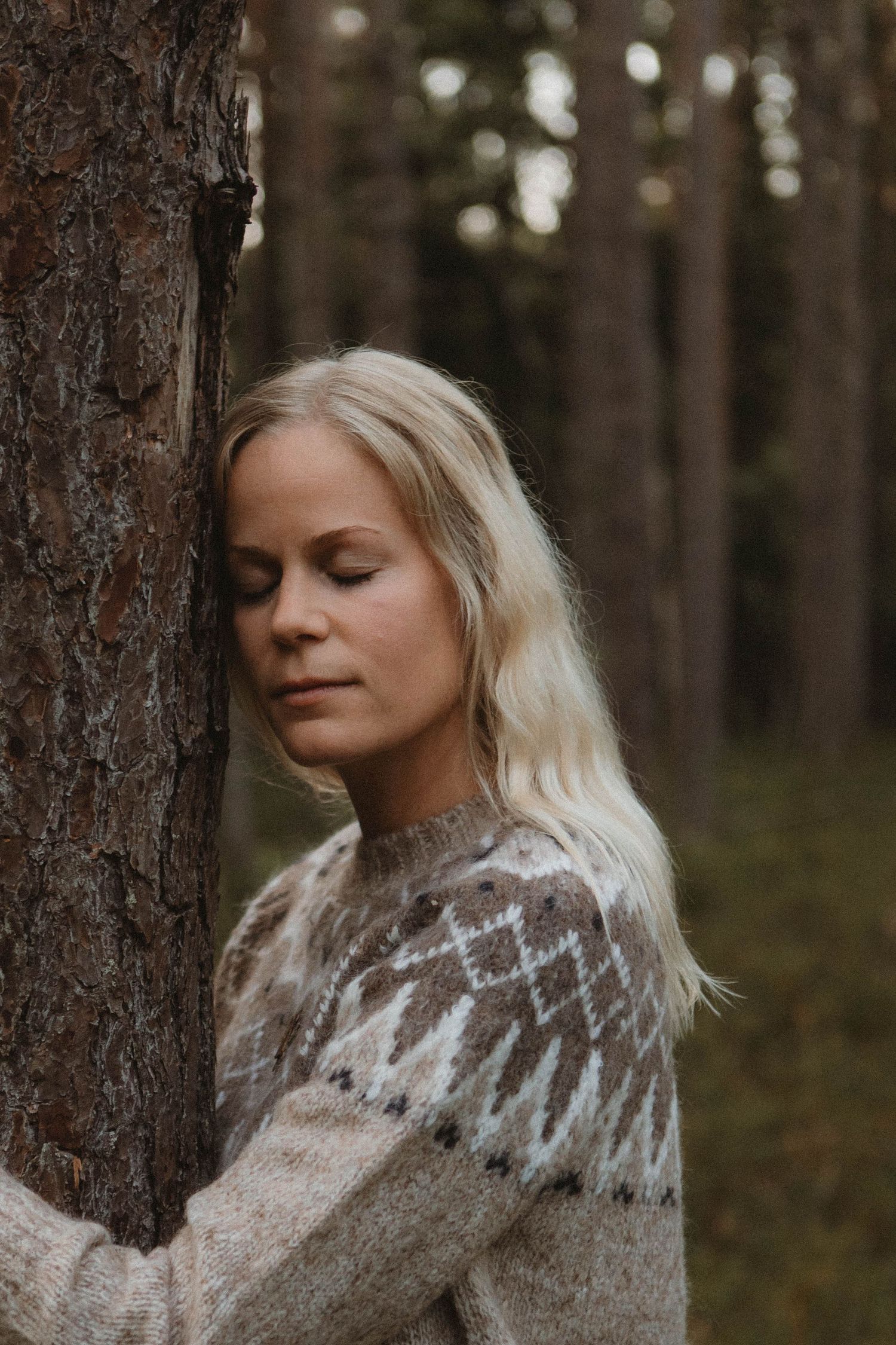 Person wearing Nordic sweater leans against tree in moody forest setting.