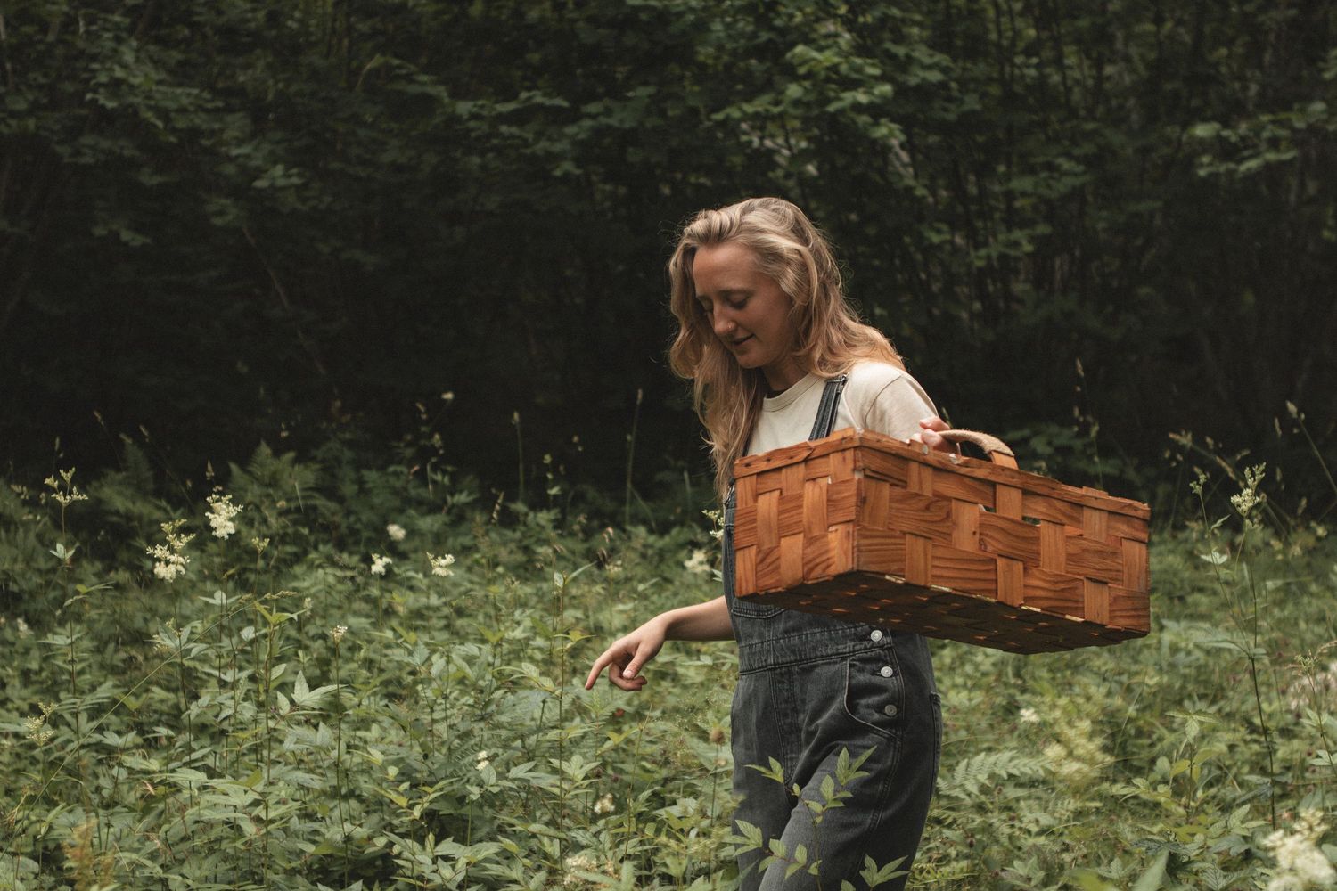 Figure walking through meadow with wooden basket in denim overalls.