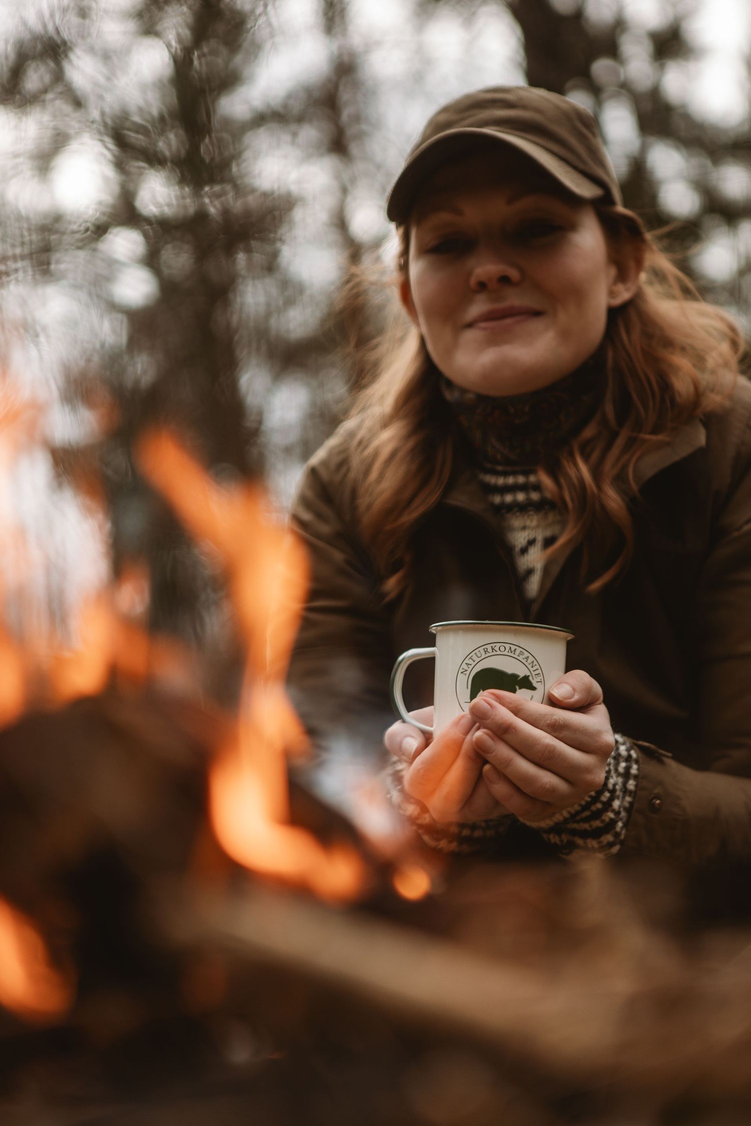 Woman holding enamel mug by a campfire in the forest, wearing a cap and brown jacket, smiling warmly.