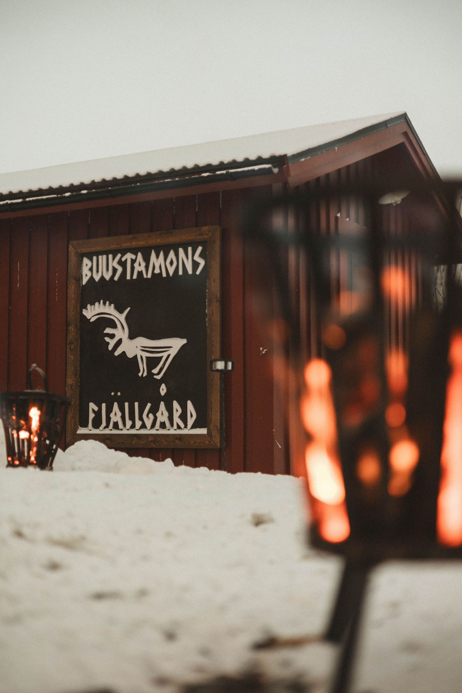 Rustic wooden cabin with 'Bundstamons' sign and moose illustration in snow with glowing lantern in foreground.