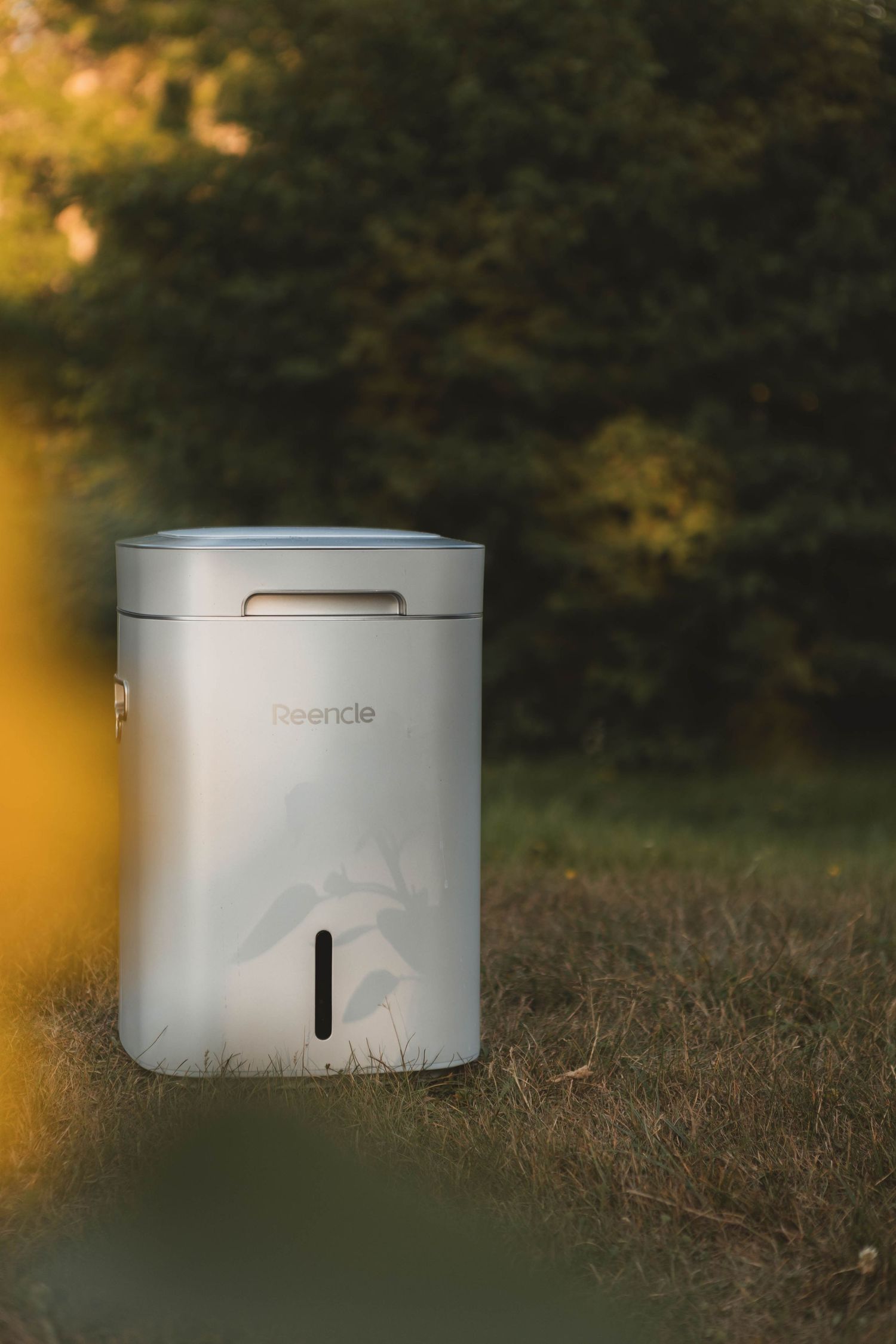 A minimalist white cylindrical smart device photographed in a natural outdoor setting with golden bokeh lighting effects.