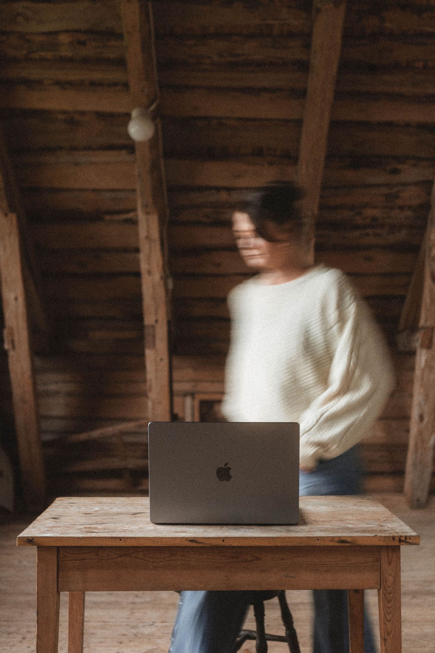 Blurred motion shot of MacBook laptop on rustic wooden table with figure in white sweater.