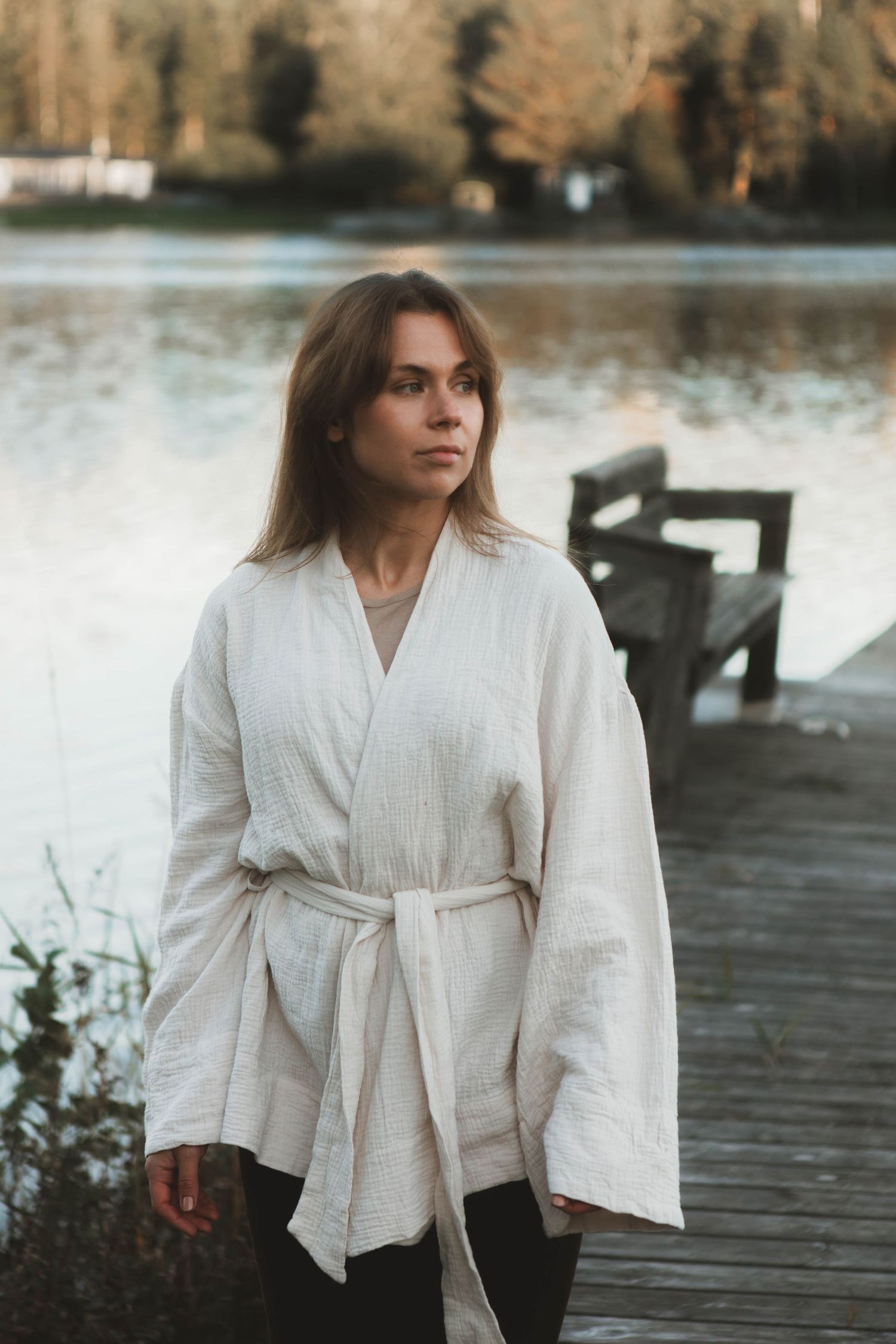 Person in flowing white kimono-style dress walking along wooden dock by tranquil lake.