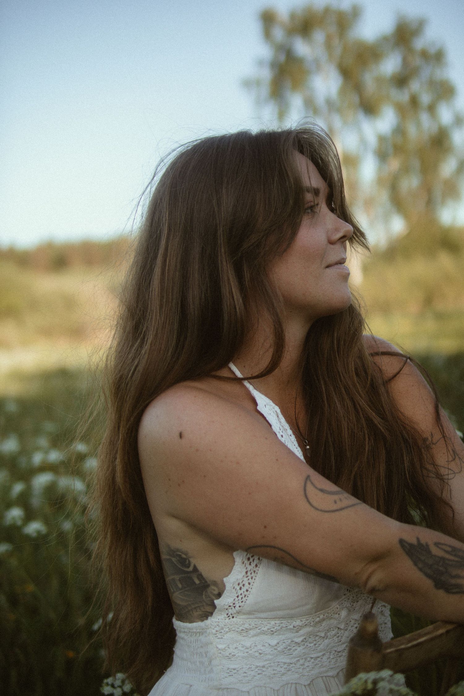 A person with flowing dark hair poses in a white dress against a natural outdoor backdrop during golden hour.