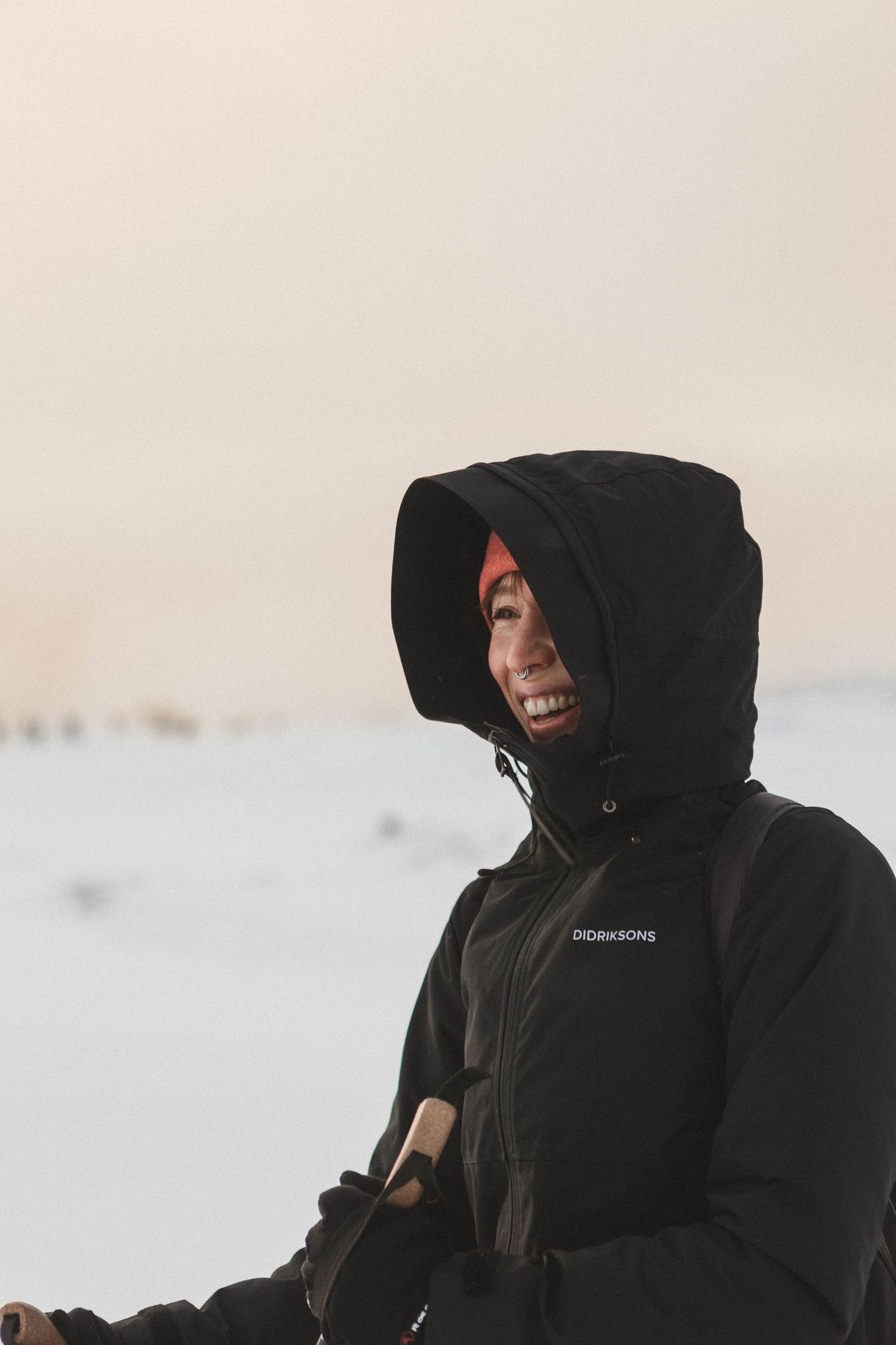 Person in a black winter coat with hood smiling while standing in a snowy landscape during winter.