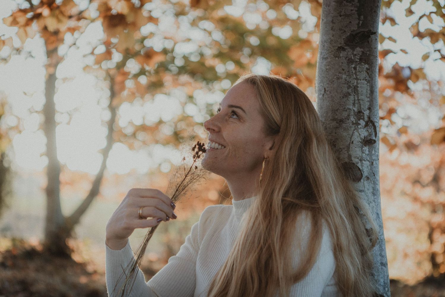 Person in white sweater leaning against tree during golden autumn sunset with soft warm lighting in background.