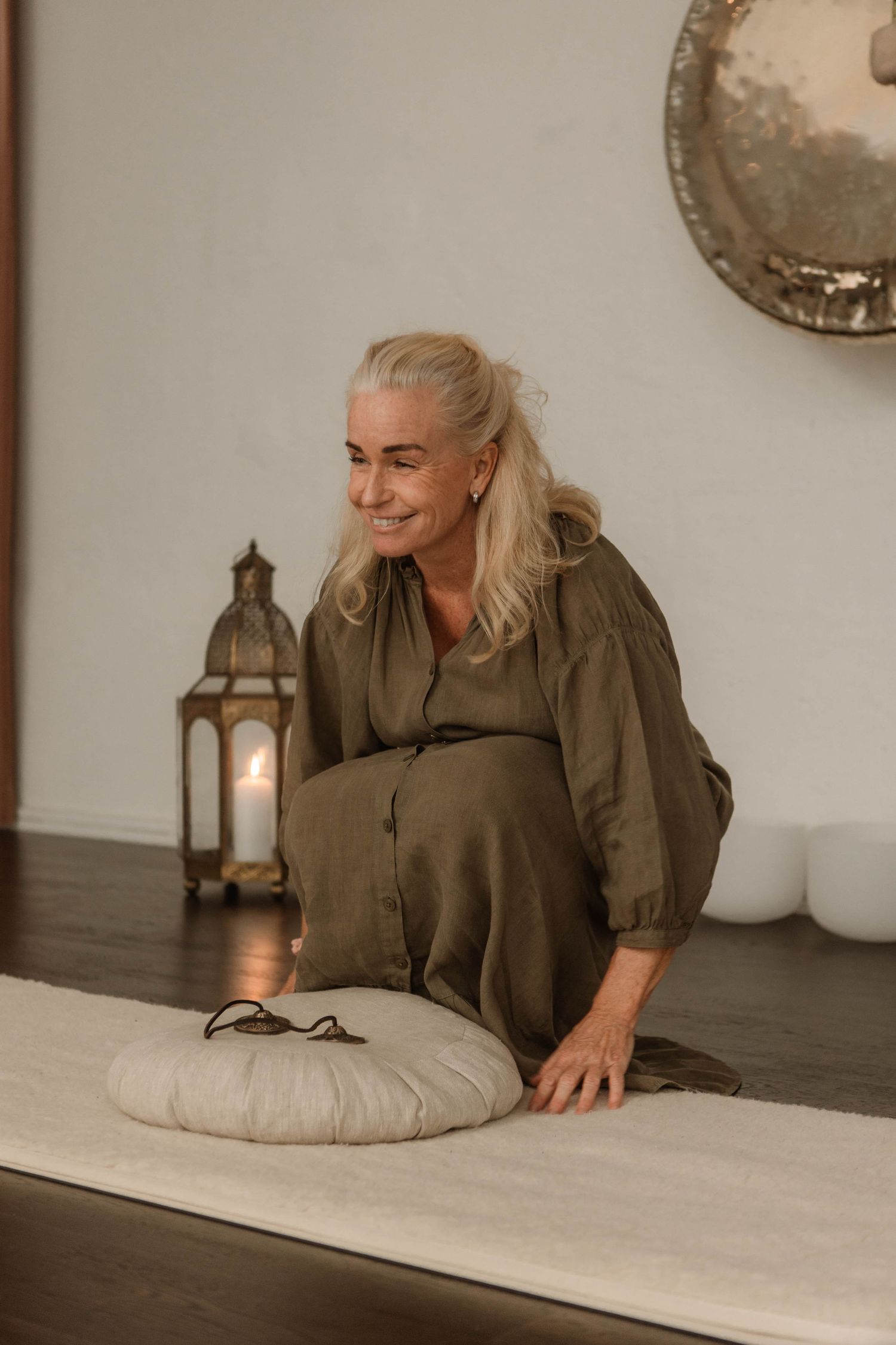 A person in olive green dress sits on floor cushions near a decorative lantern in a warmly lit room.