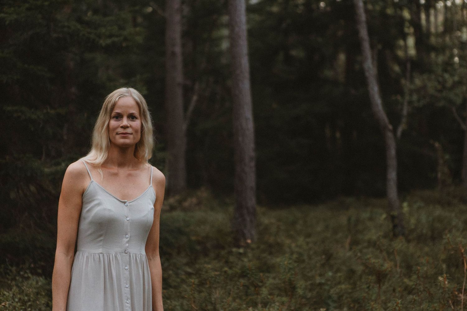 Figure in simple grey dress stands among tall pine trees in atmospheric forest scene.