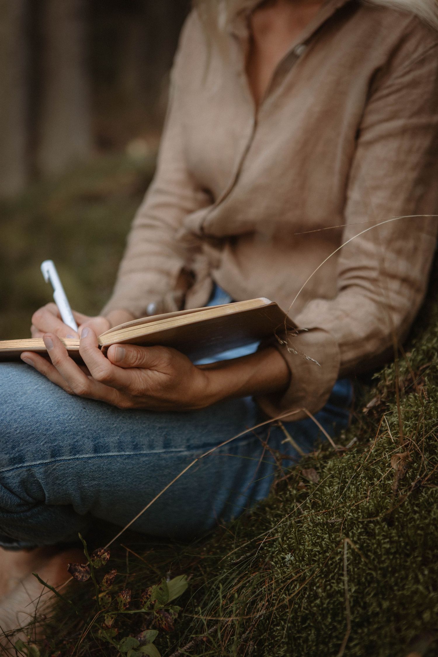 Close-up of hands writing in notebook while sitting on mossy ground wearing jeans.