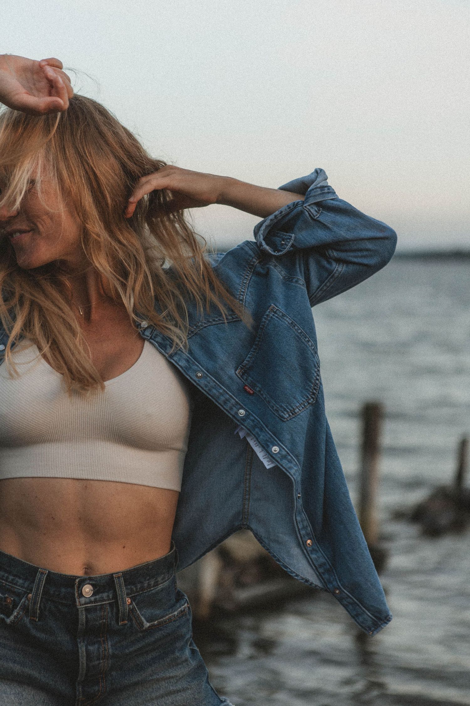 Woman in crop top and denim jacket dances by the ocean at sunset.