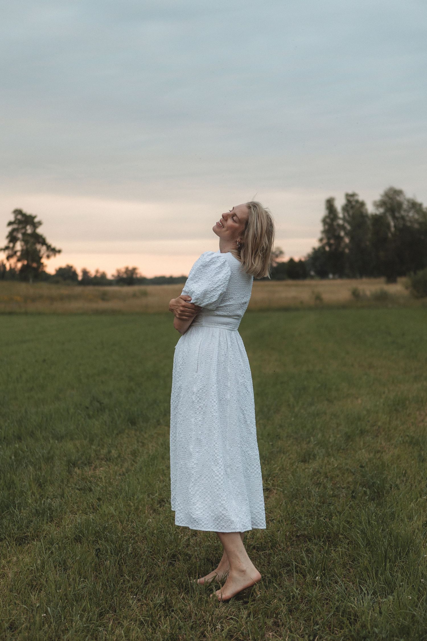 Someone in a white dress stands barefoot in a field during golden hour.