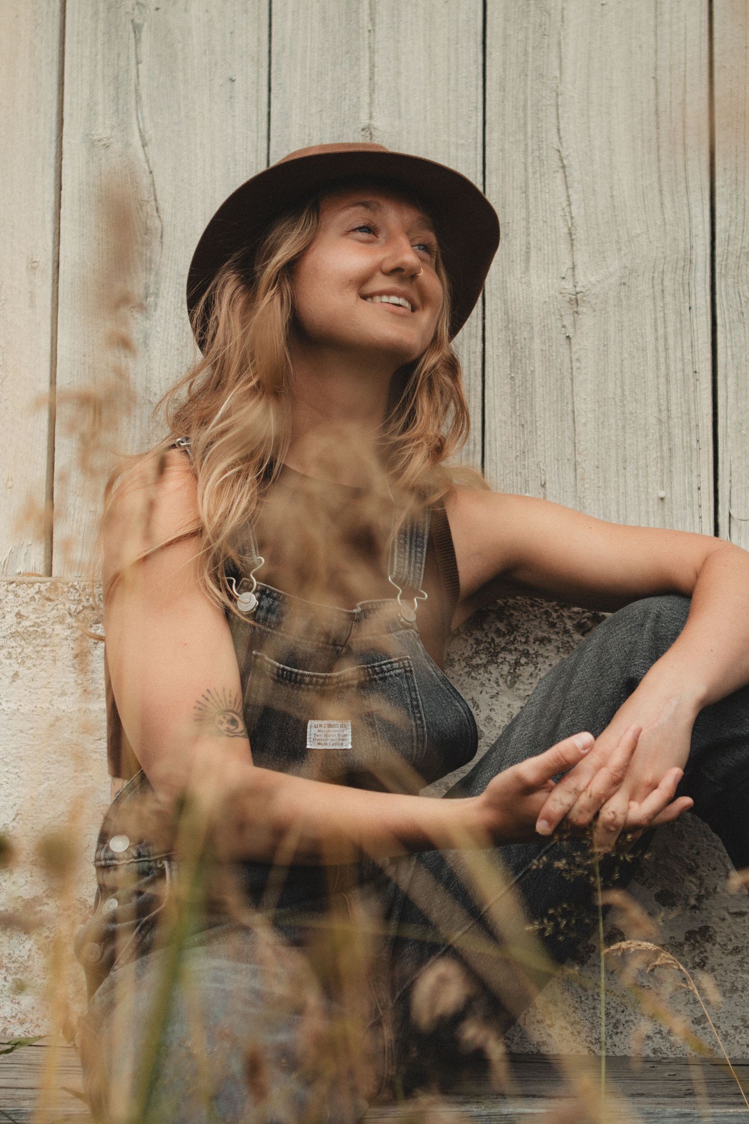 Woman in denim overalls and hat sits casually against rustic wooden wall in sunlit outdoor setting.