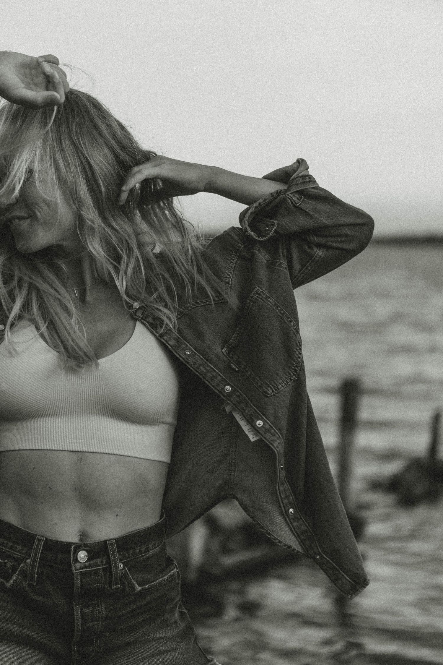 Person in denim jacket and crop top with windswept hair against ocean backdrop.