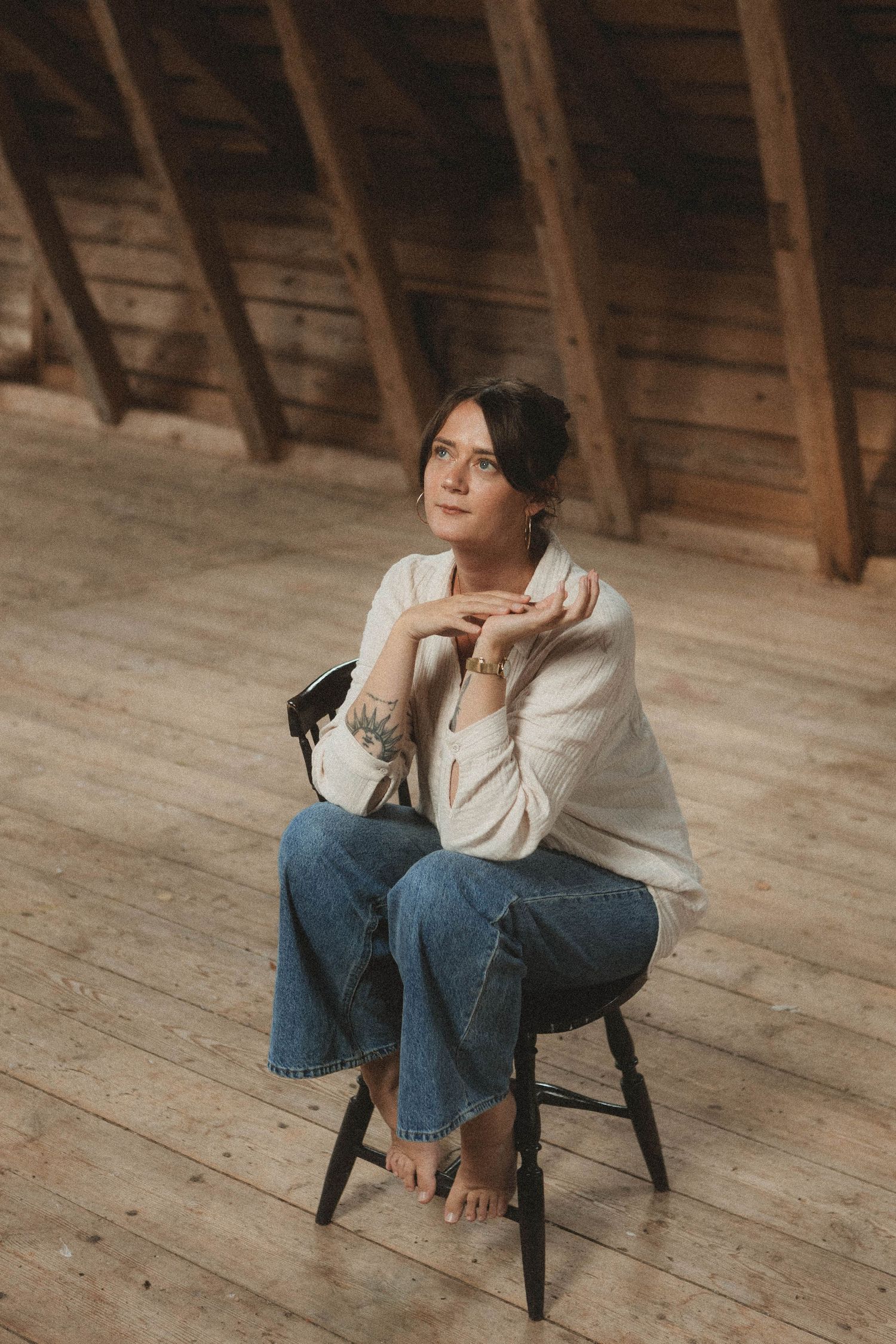 Person in casual attire sitting thoughtfully on dark chair in wooden attic space.
