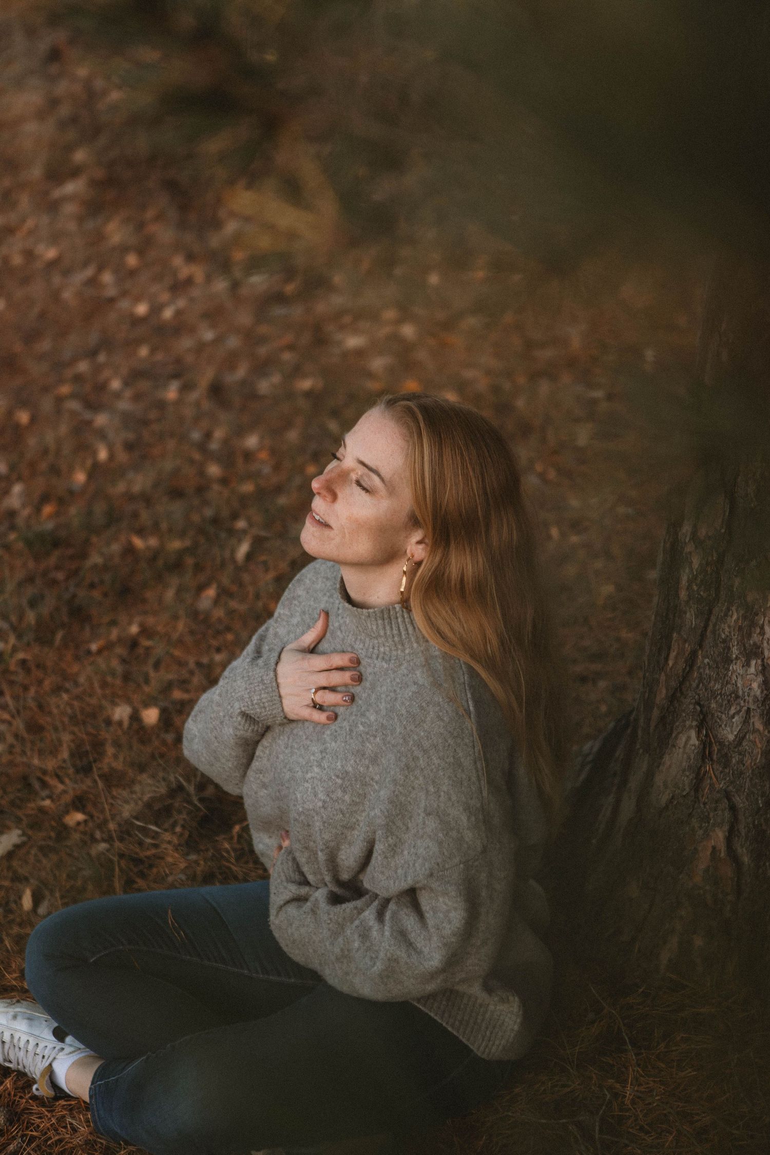 A person in a cozy gray sweater sits against a tree trunk in an autumn forest setting with fallen leaves.