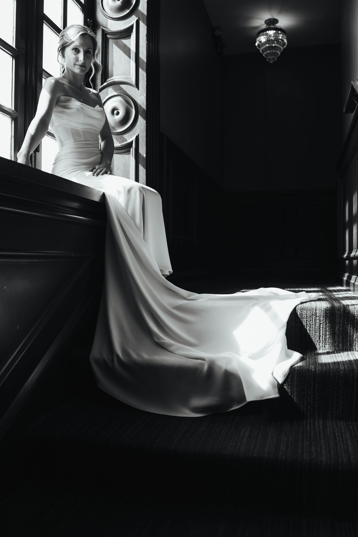 Elegant black and white bridal portrait showing flowing white wedding gown cascading down dark stairs near ornate window.