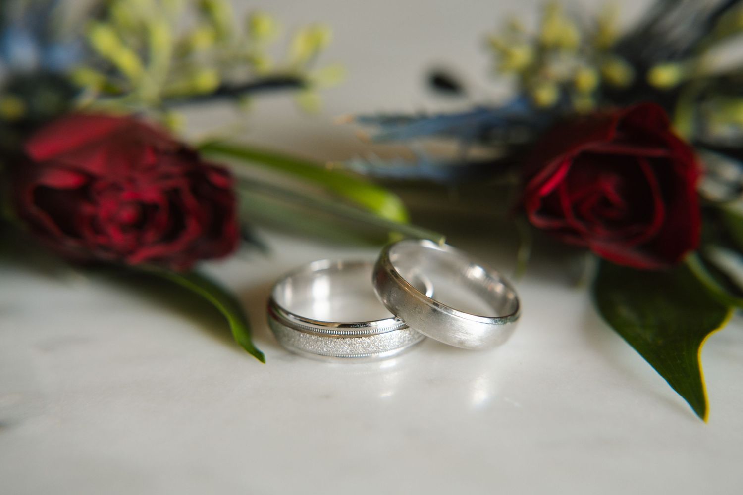 Two silver wedding rings on a white surface with red roses in the background.