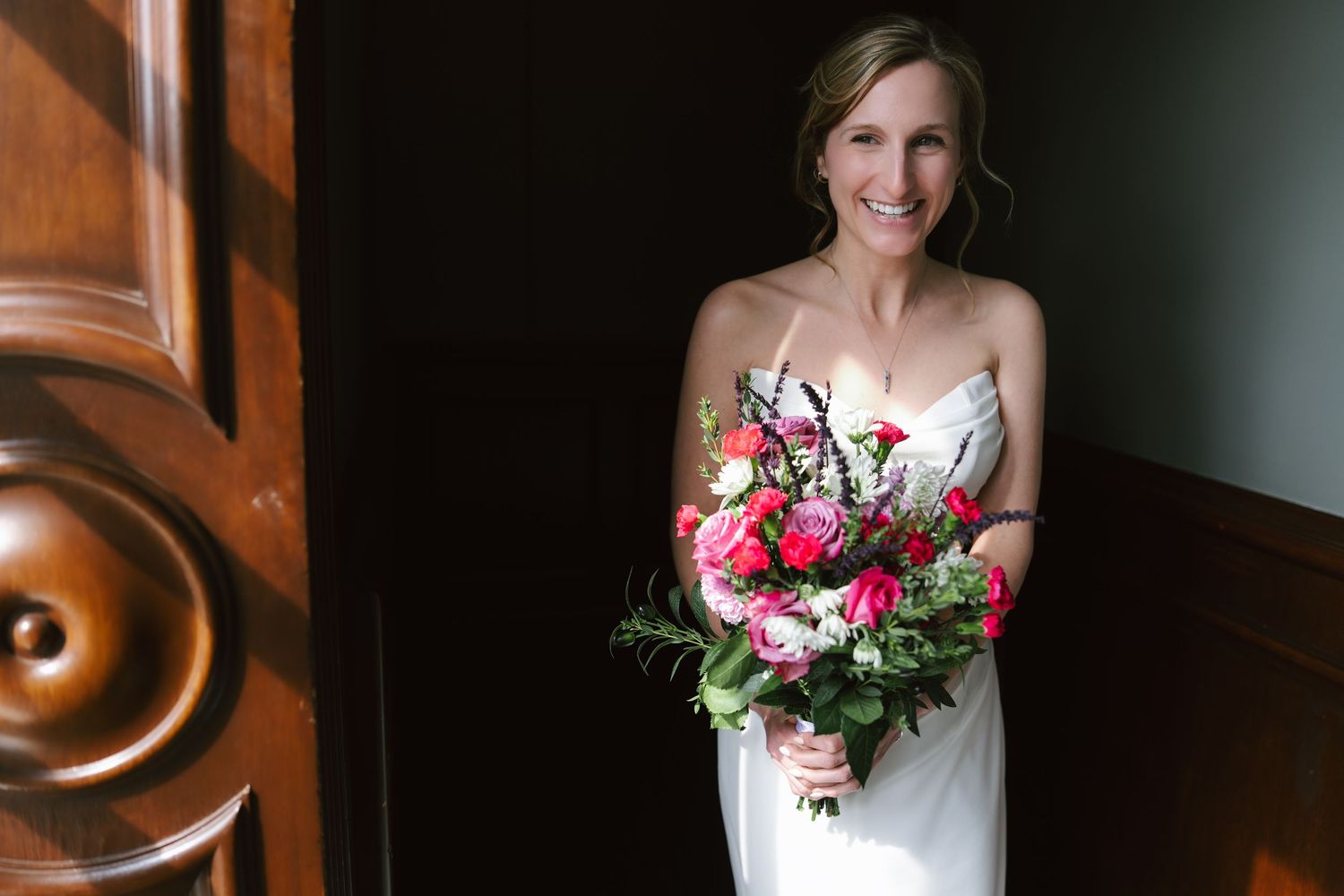 A bride in a white strapless gown holds a vibrant pink and red floral bouquet while standing against a dark wall.
