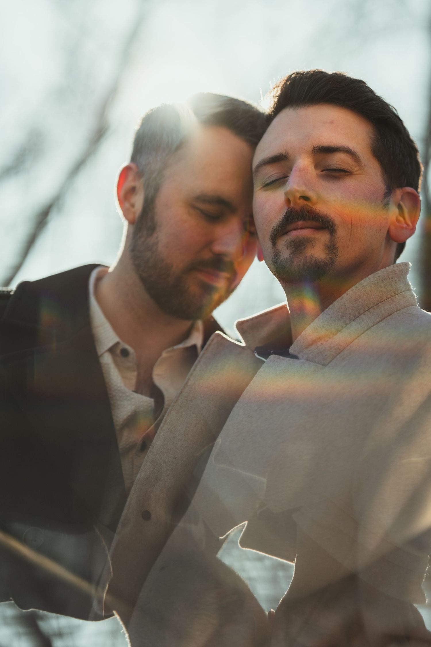 Two men in suits share a romantic moment outdoors, bathed in golden sunlight with a colorful lens flare.