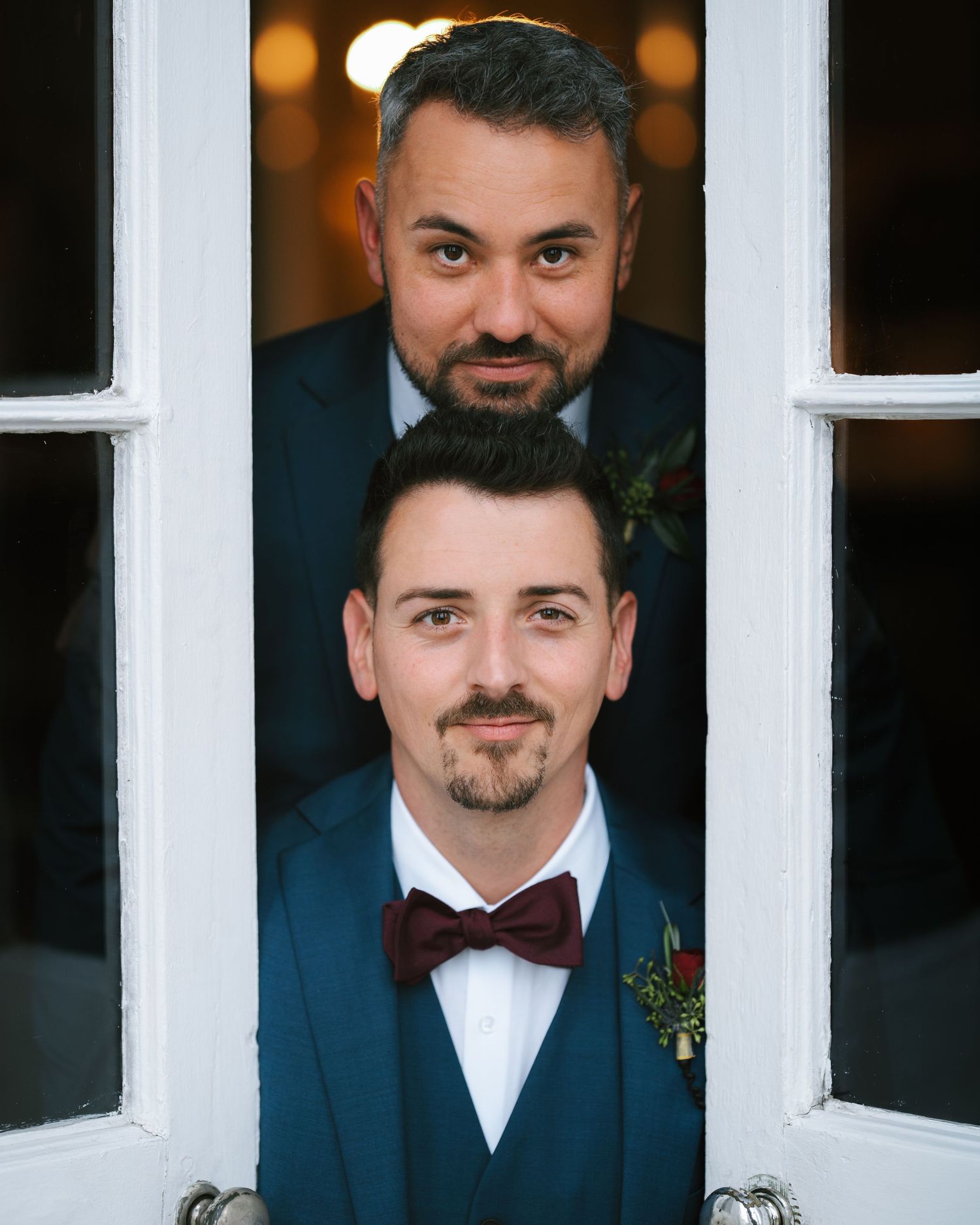 Two men in formal attire, one with a beard and the other clean-shaven, stand close together and look directly at the camera.