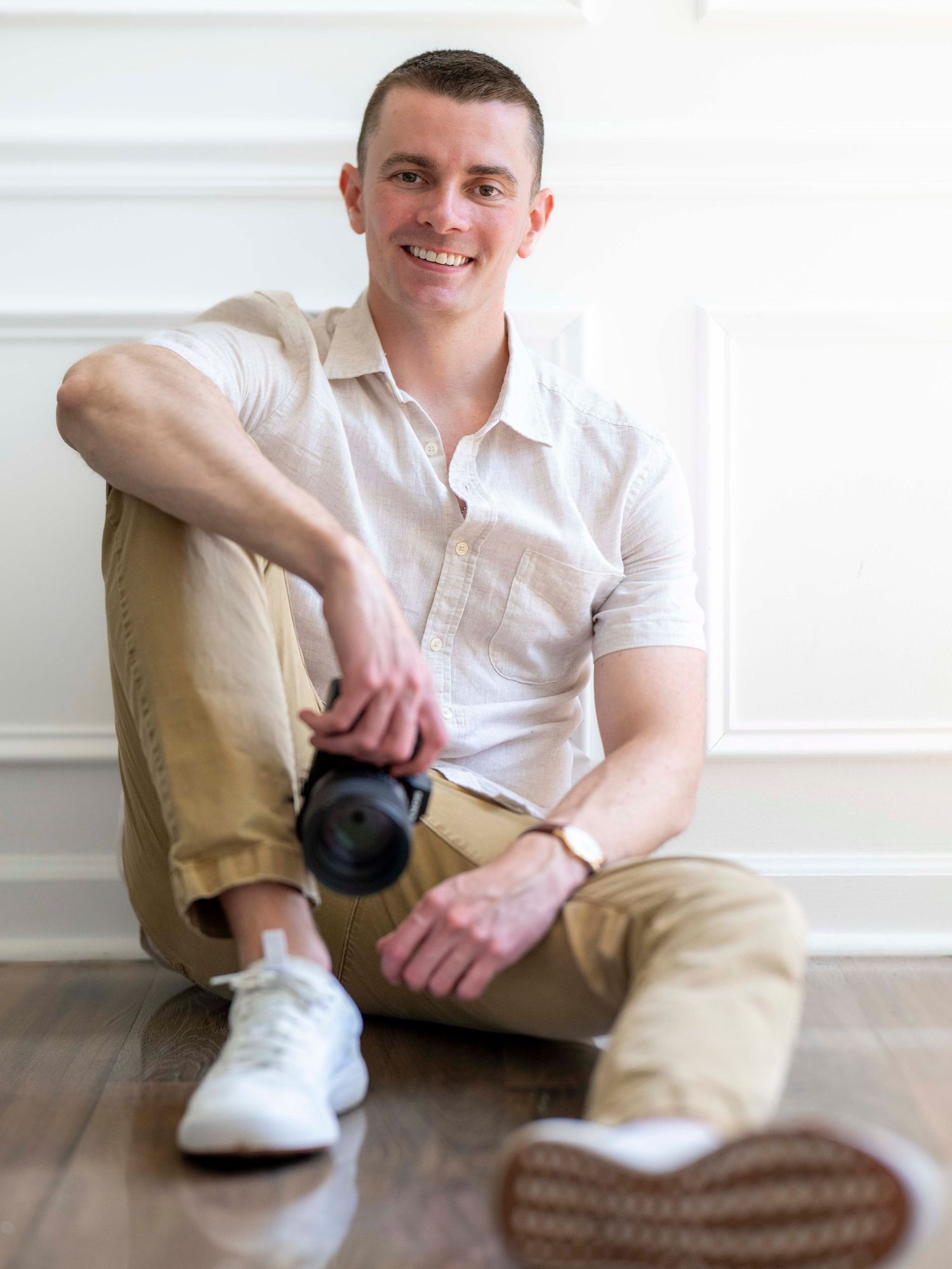 A person in a white polo shirt and khaki pants sits casually on the floor against a light-colored wall.
