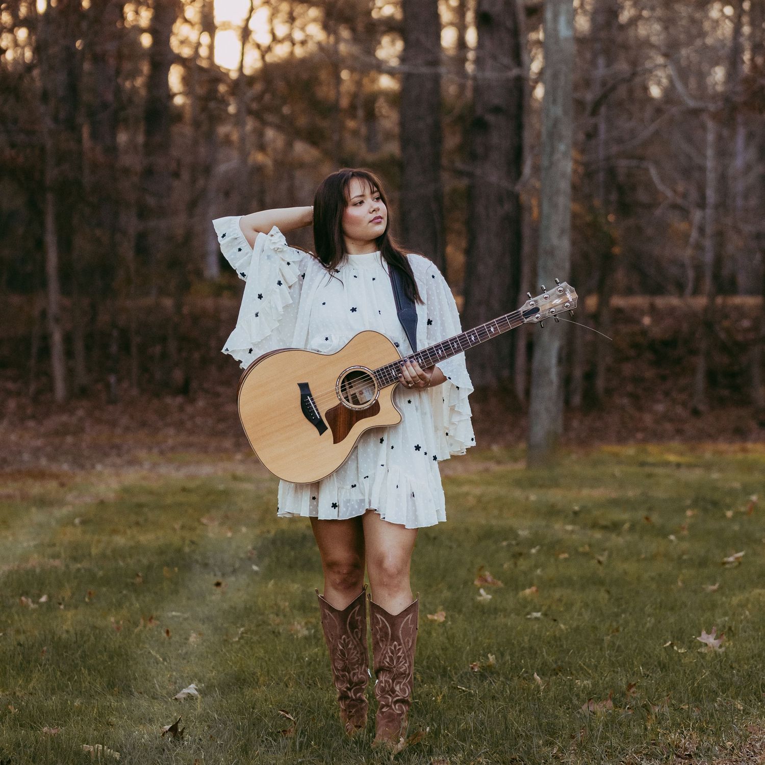 A person in a white polka dot dress holds an acoustic guitar in a wooded outdoor setting at sunset.