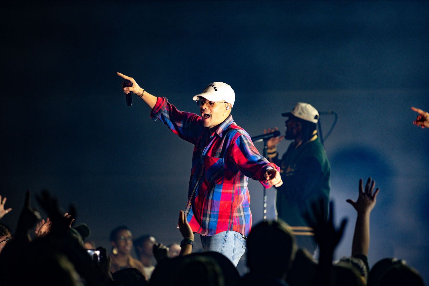 Concert performer in a baseball cap and plaid shirt gestures to the crowd during a high-energy performance on stage.
