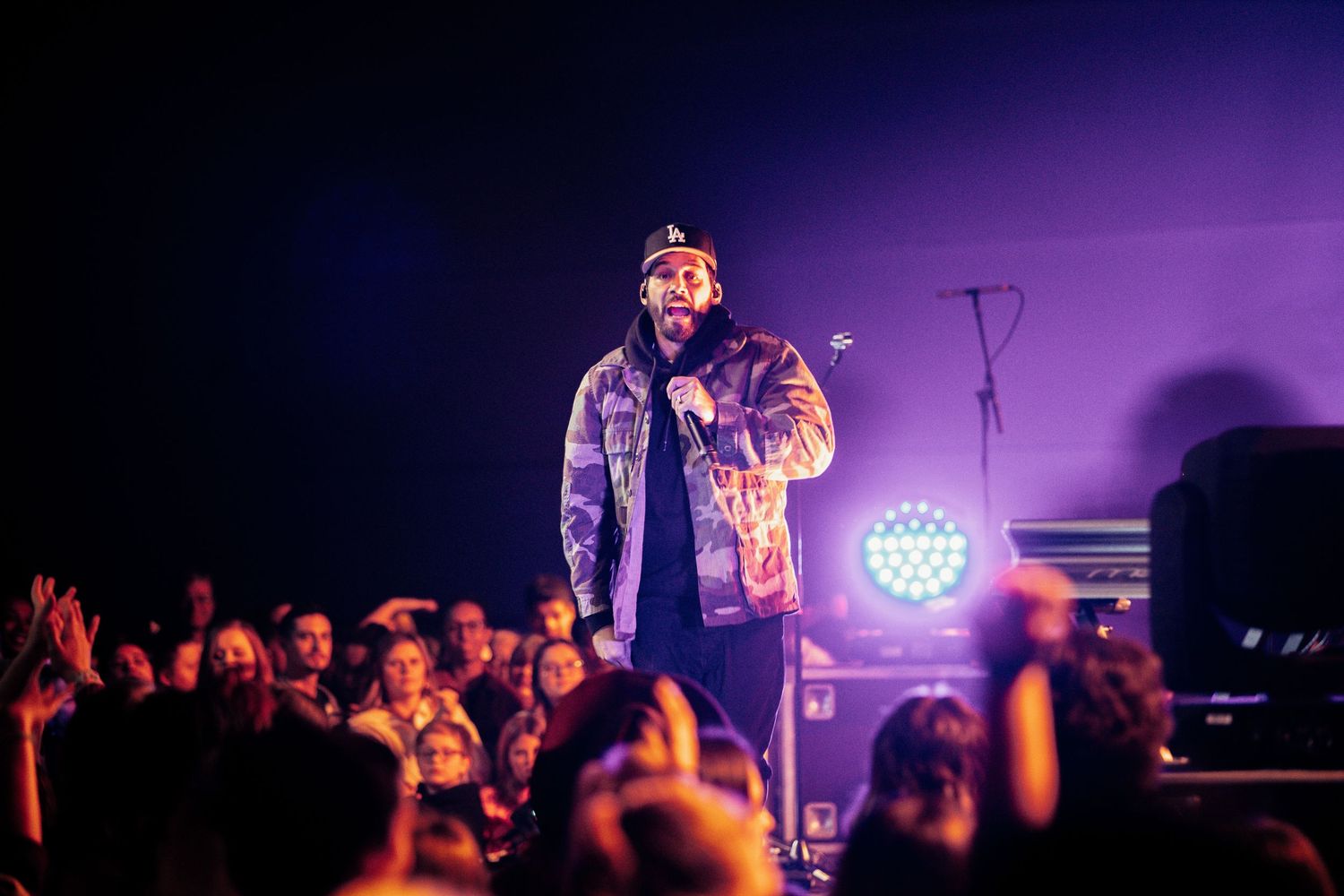 A performer stands on stage in denim and camo jacket illuminated by purple lighting while crowd watches in silhouette.