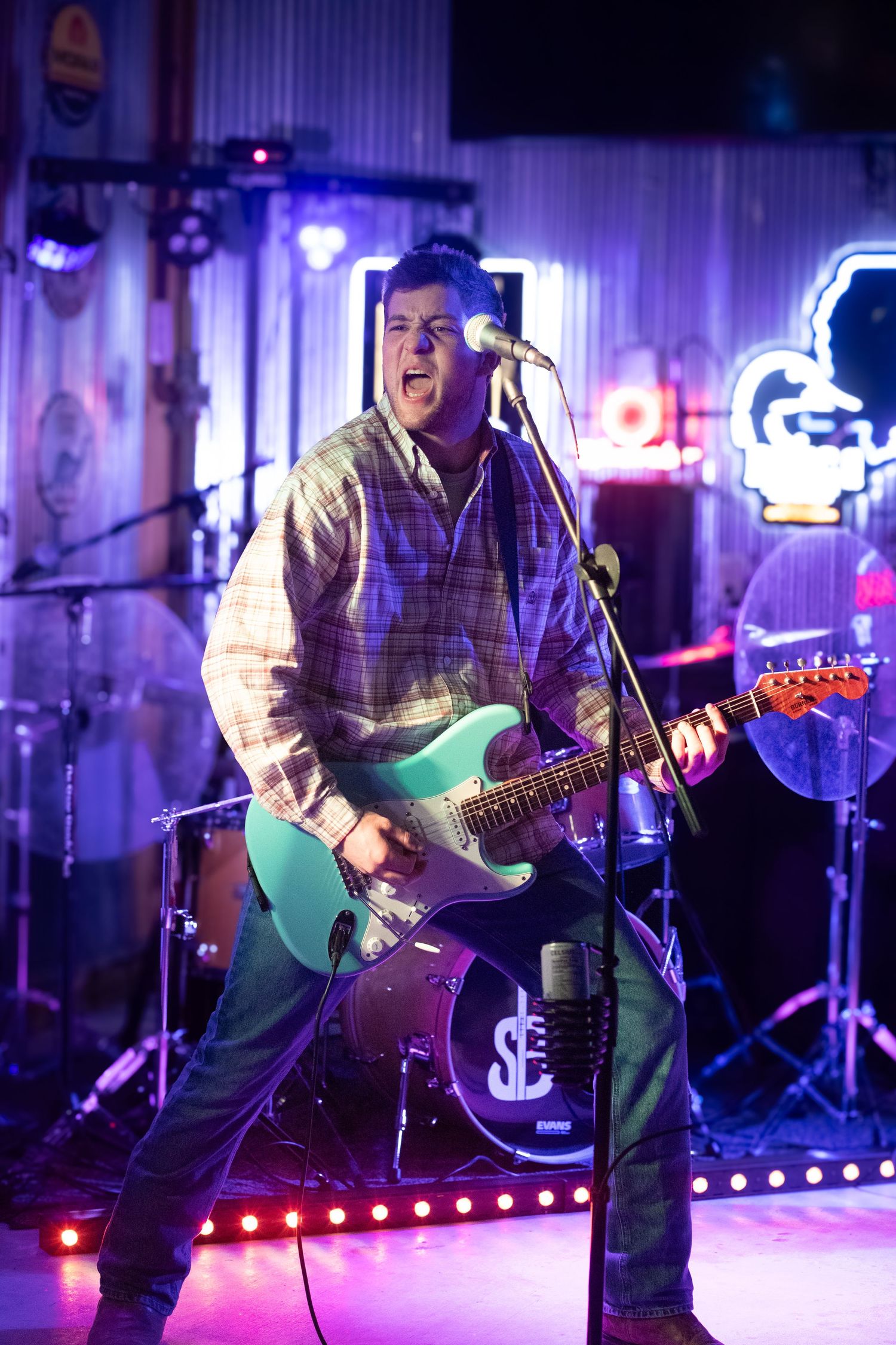 Musician performs energetically on stage with guitar under purple and blue lighting during live concert performance.