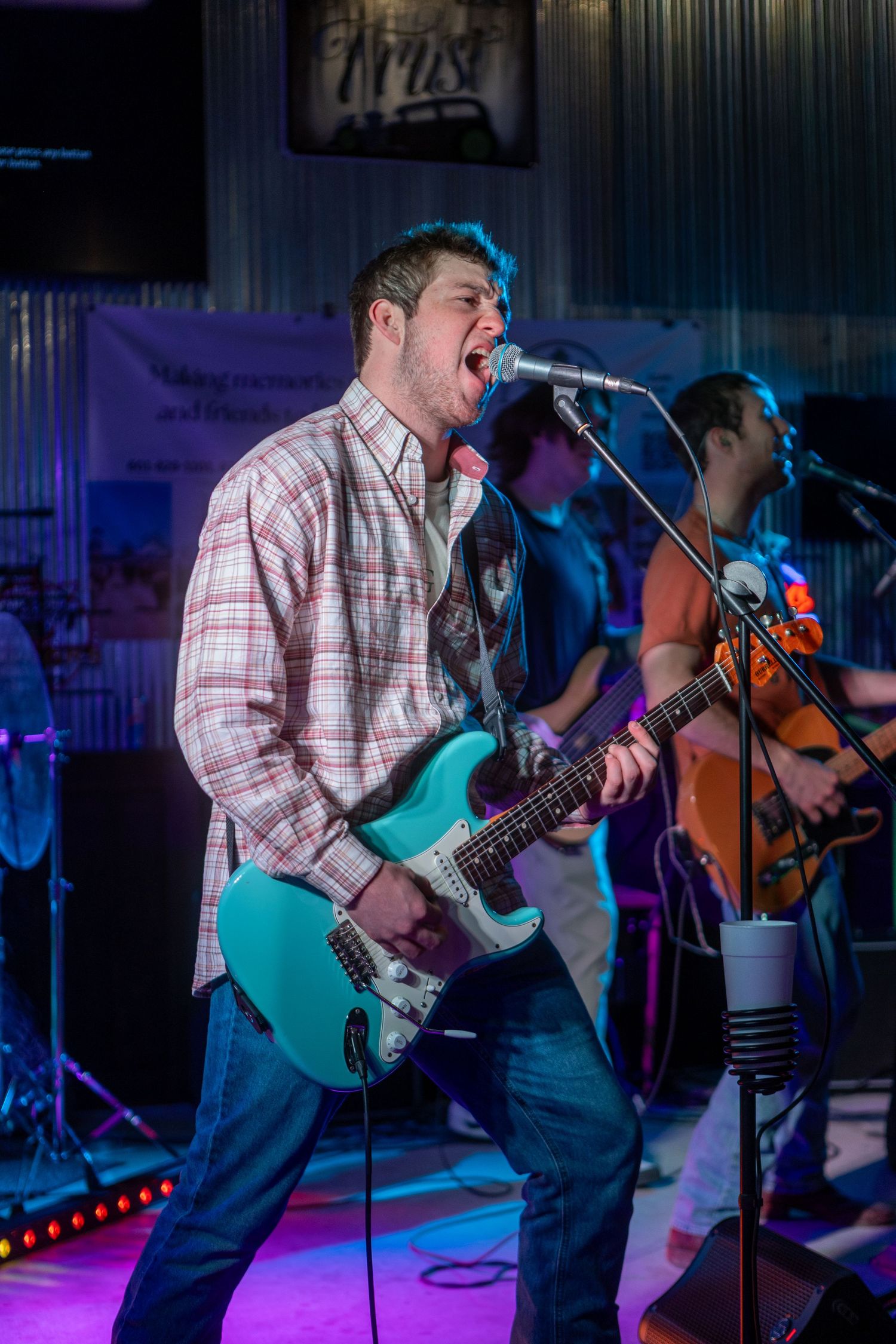 Male guitarist in plaid shirt singing passionately into microphone on stage with band performing behind him.