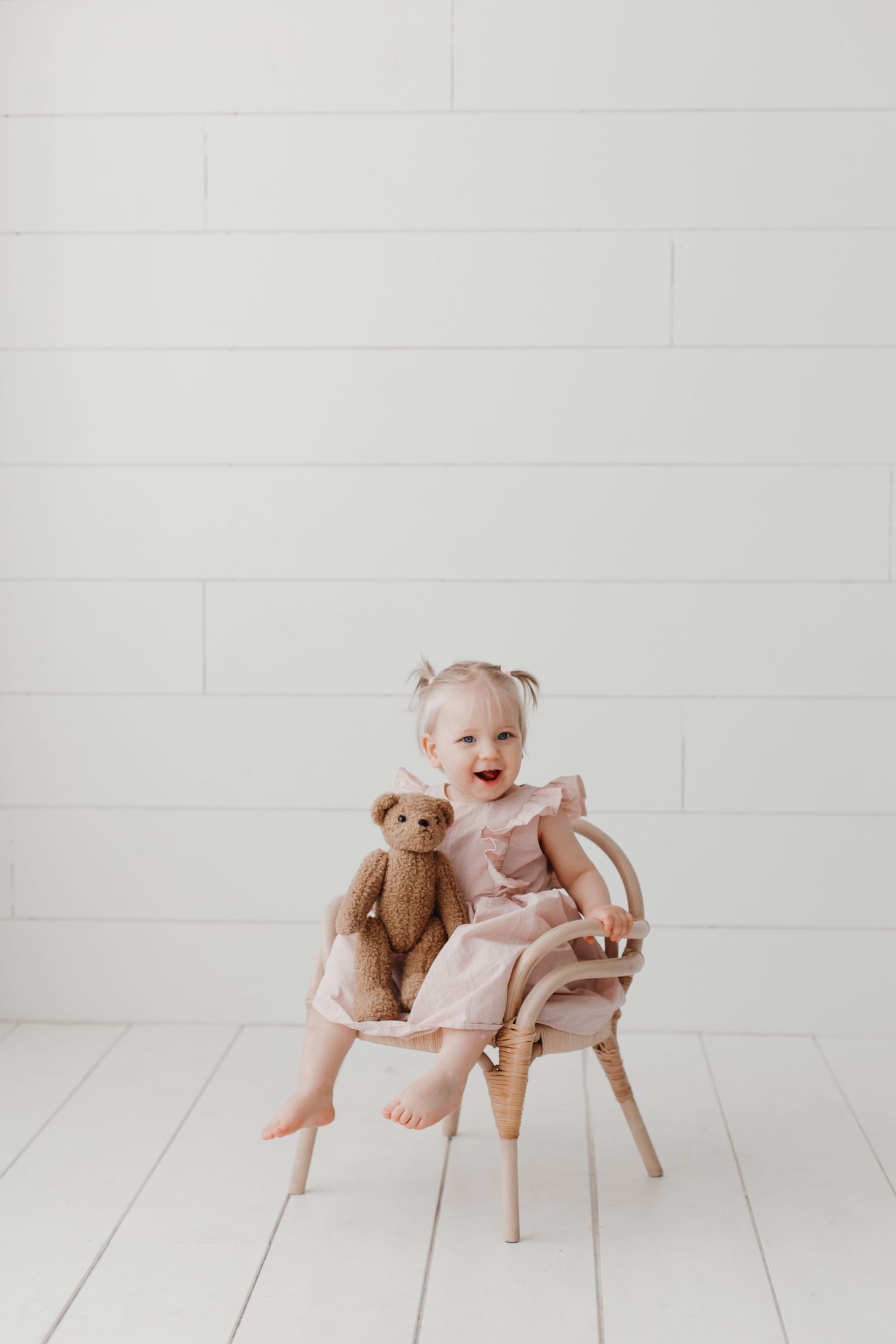 A small child sits in a wooden chair with a teddy bear in a minimalist white room.