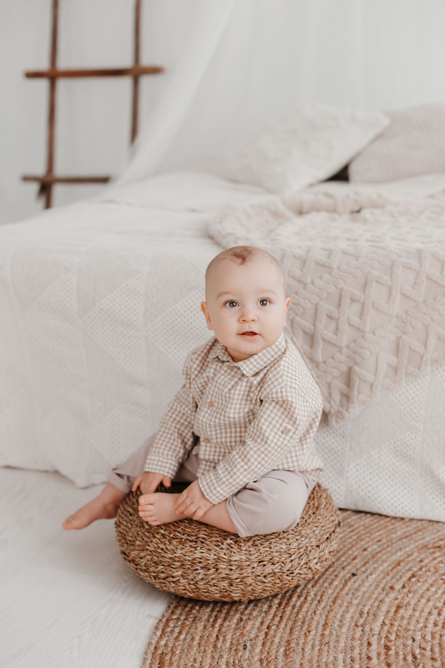 Baby in checkered shirt sitting in woven basket against textured bedding and minimal decor.