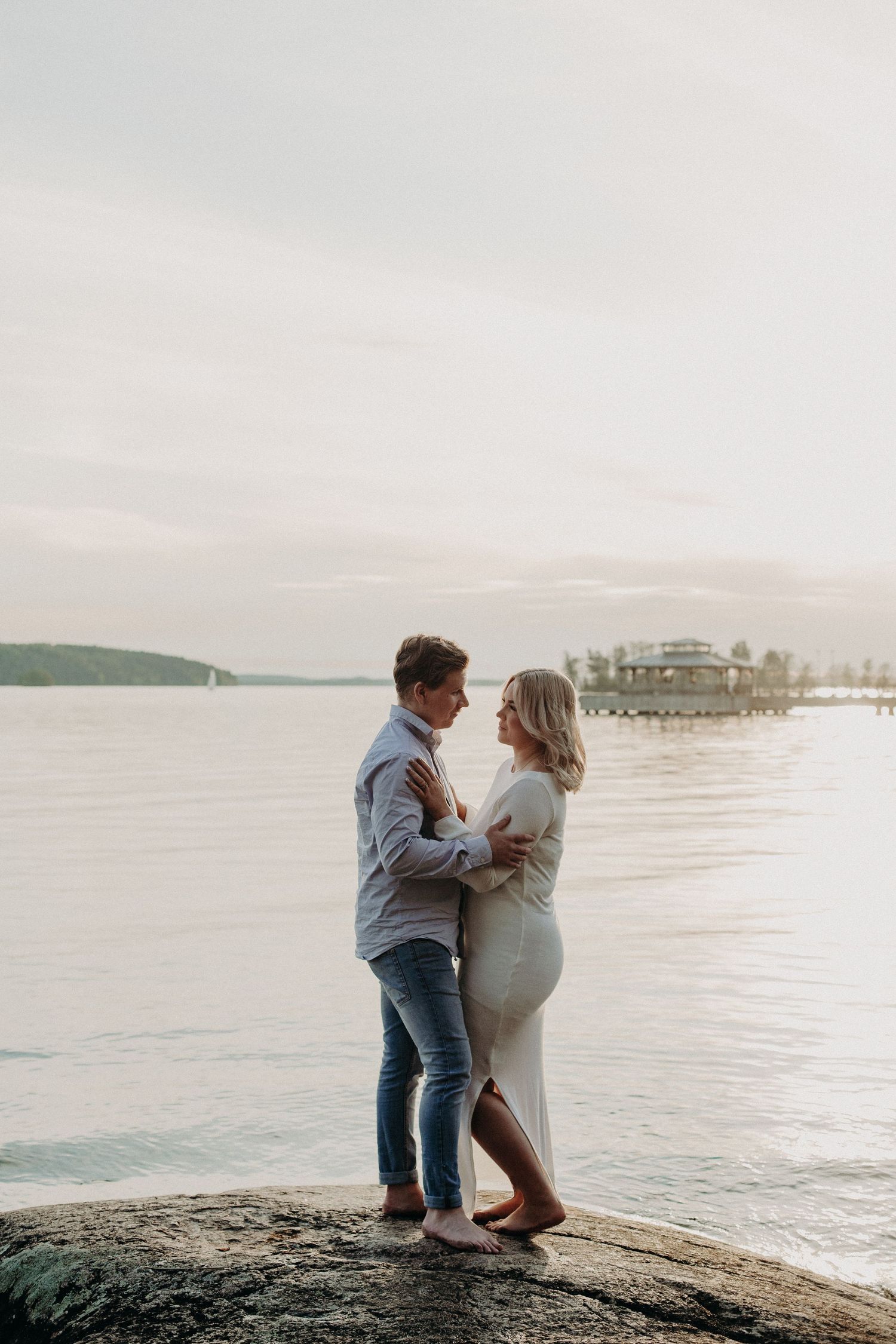 Couple facing each other on rocky shoreline at sunset, embracing with calm water and pier in background.