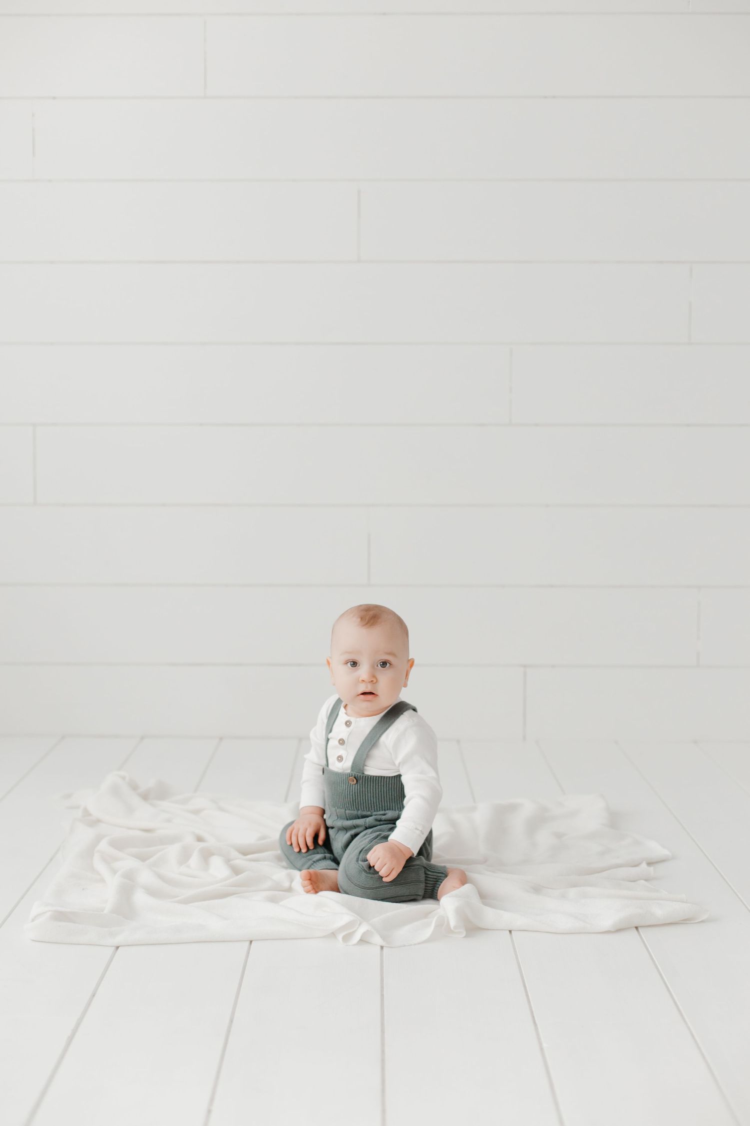 Baby in grey overalls sitting on white wooden floor with blanket.