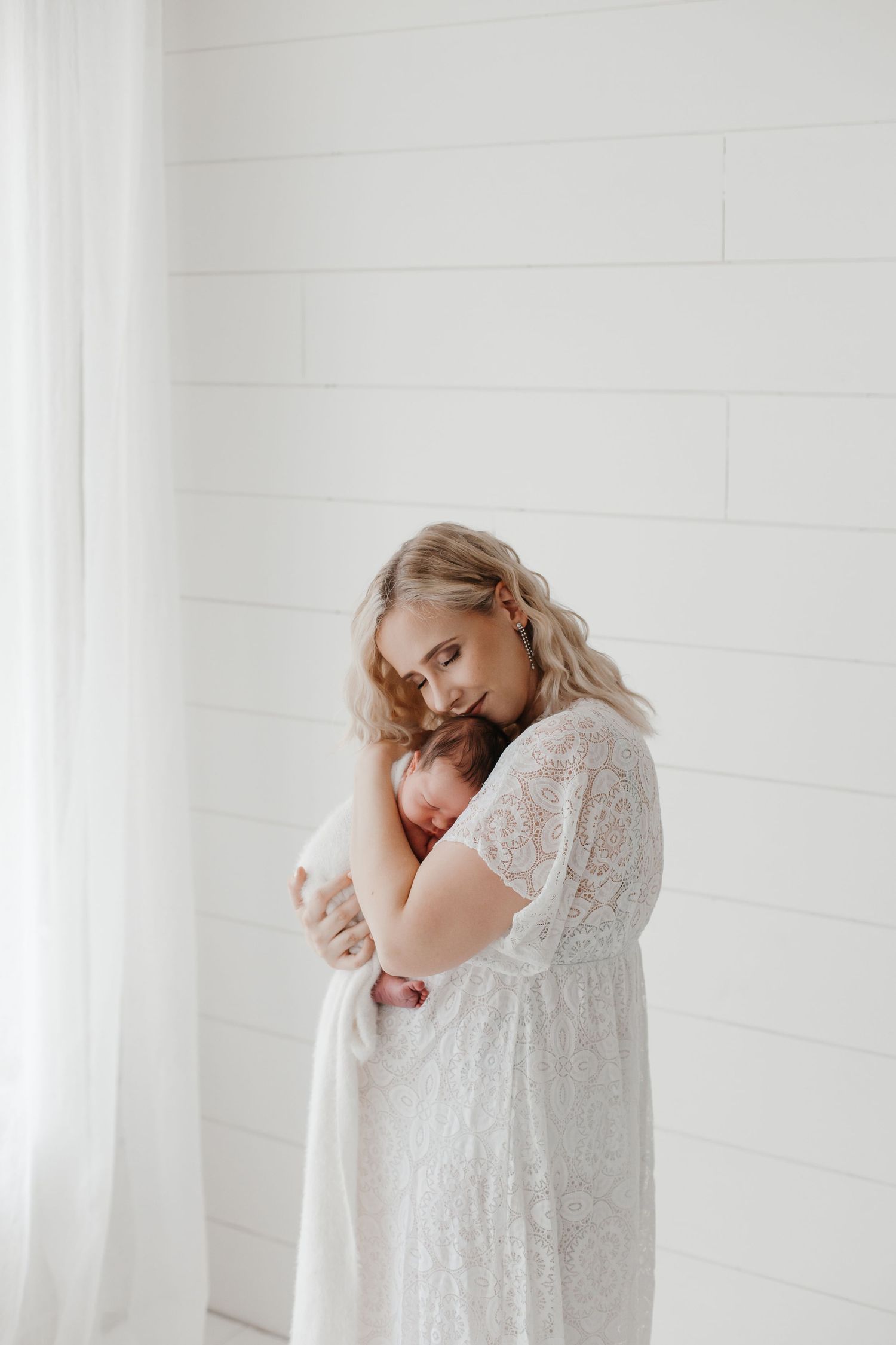 Mother in delicate white dress holding baby in bright studio setting.