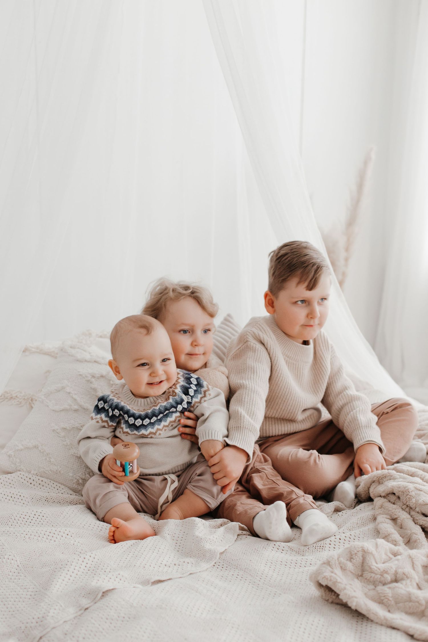 Babies in neutral colored outfits sitting together on white bedding.