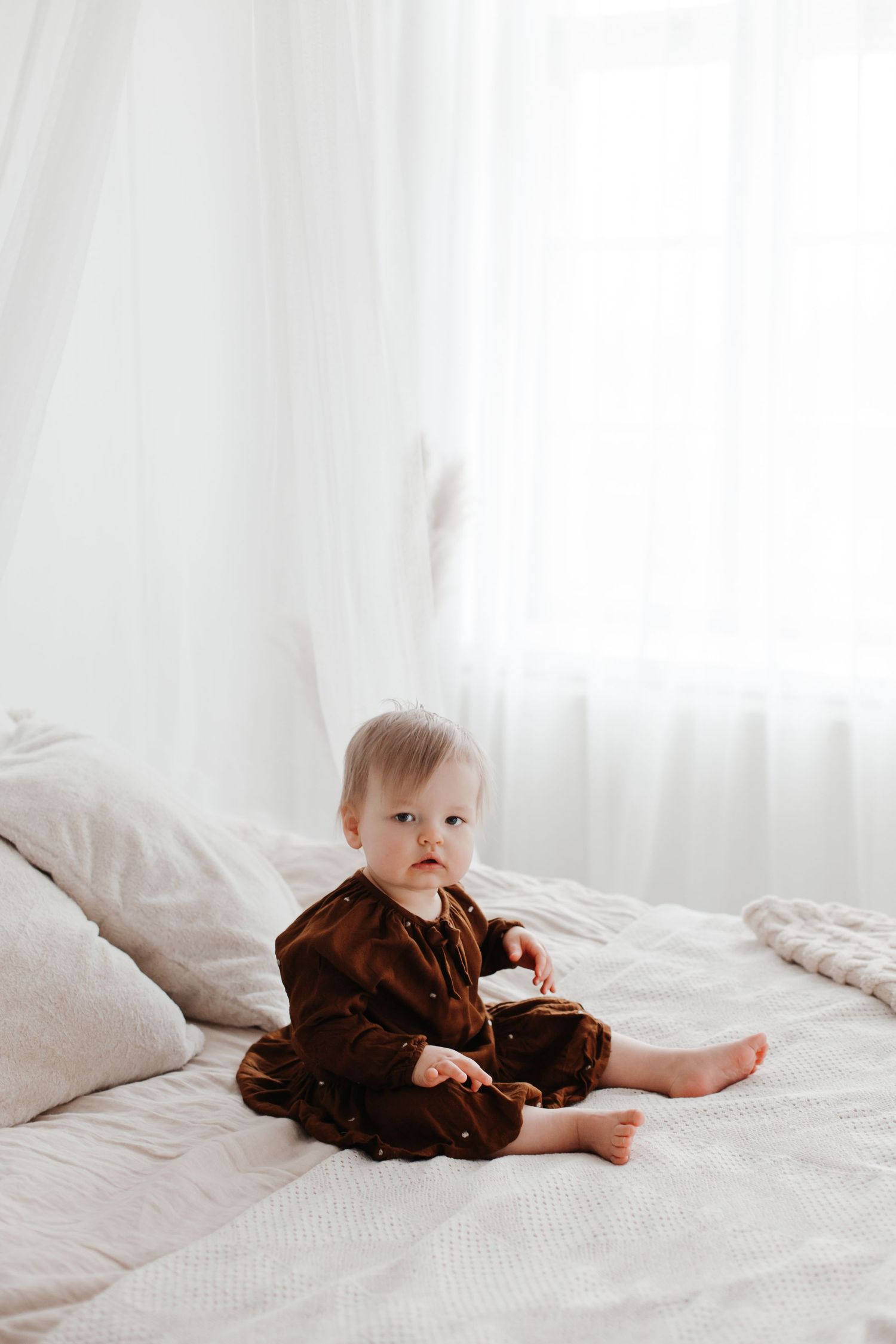 Smiling baby in brown outfit sitting on white bed.