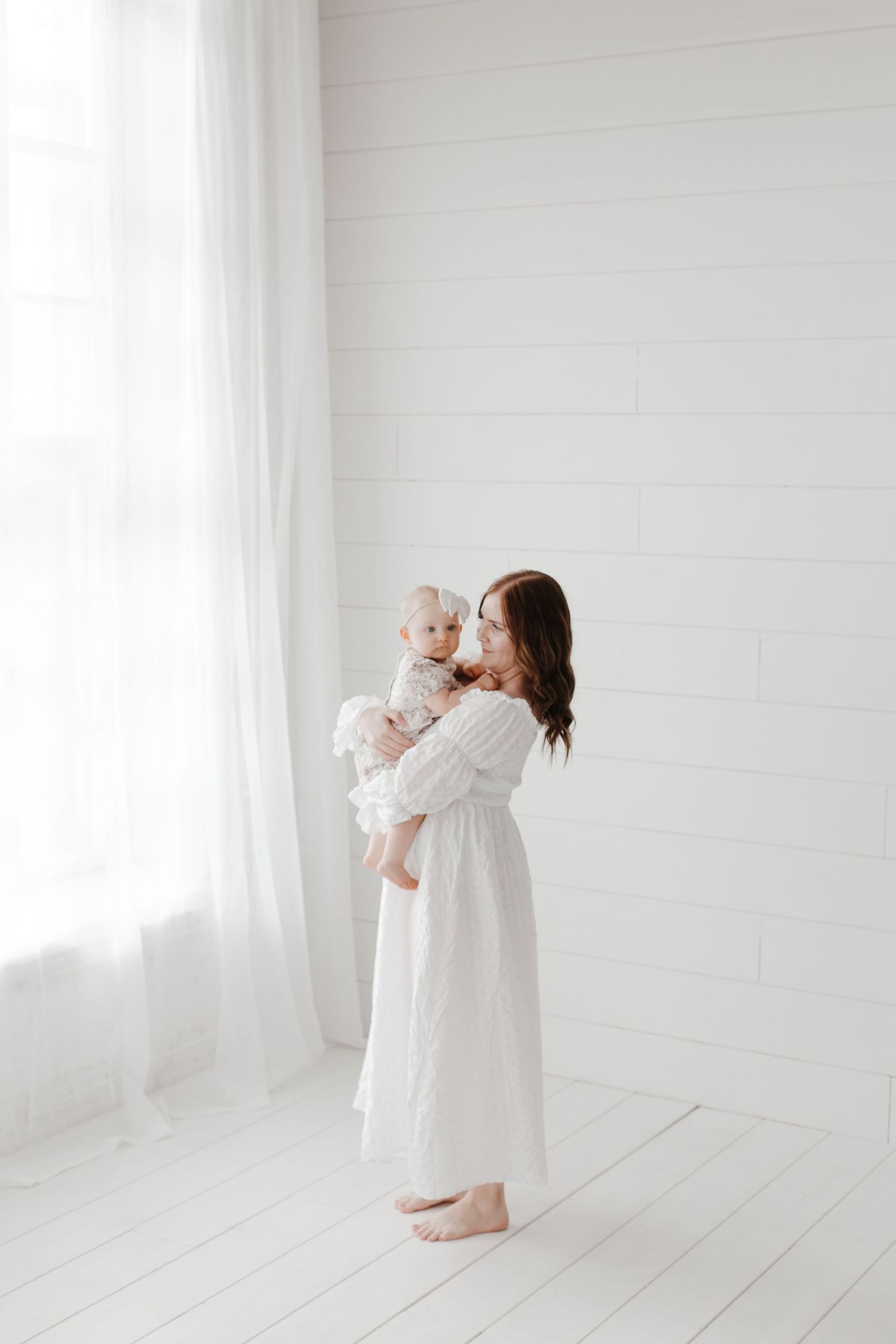 Mother and baby in white dresses share loving moments against white backdrop.