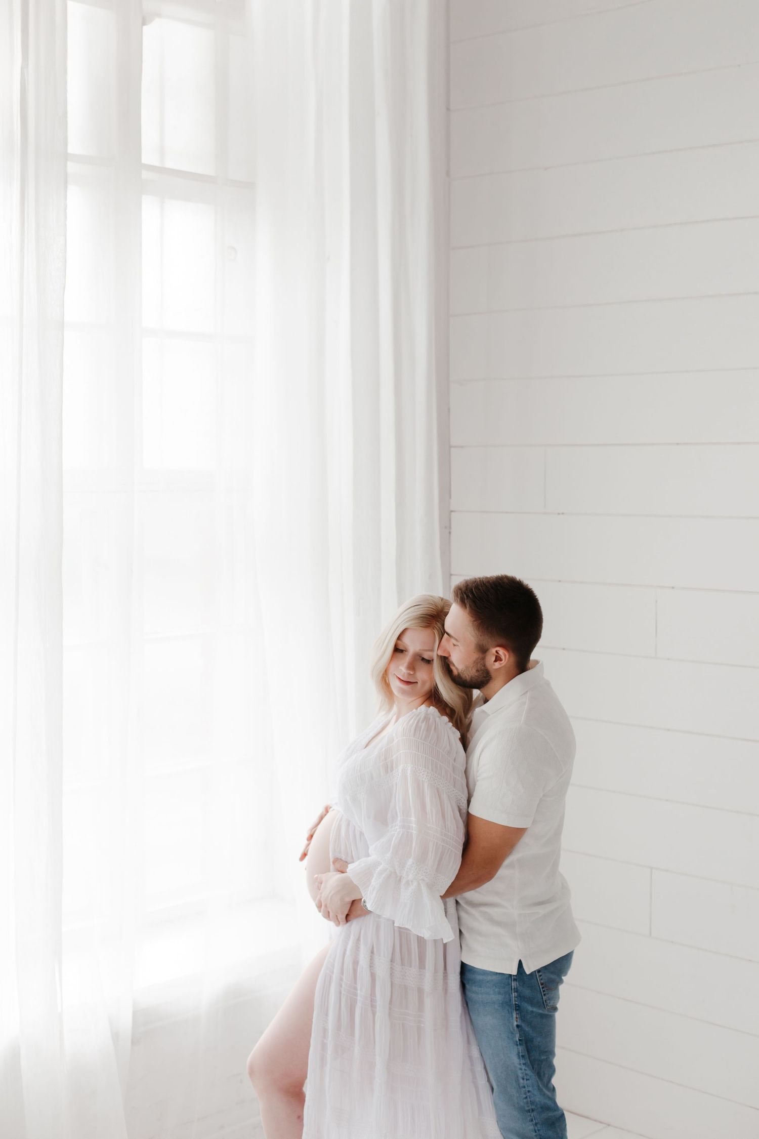 Romantic maternity portrait against white wall with soft window light.