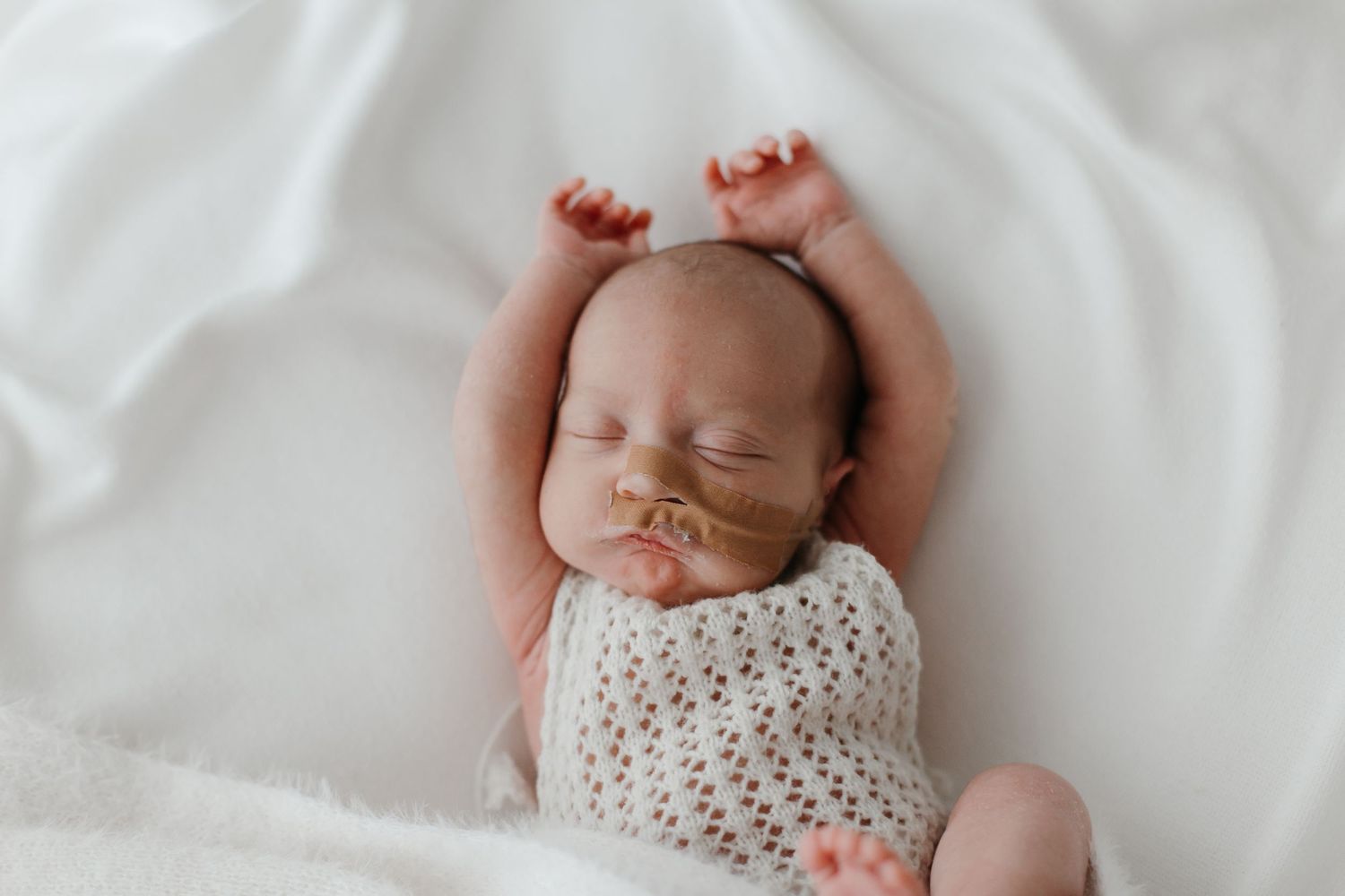 Sleeping newborn baby wearing a delicate white knit outfit against soft white bedding.
